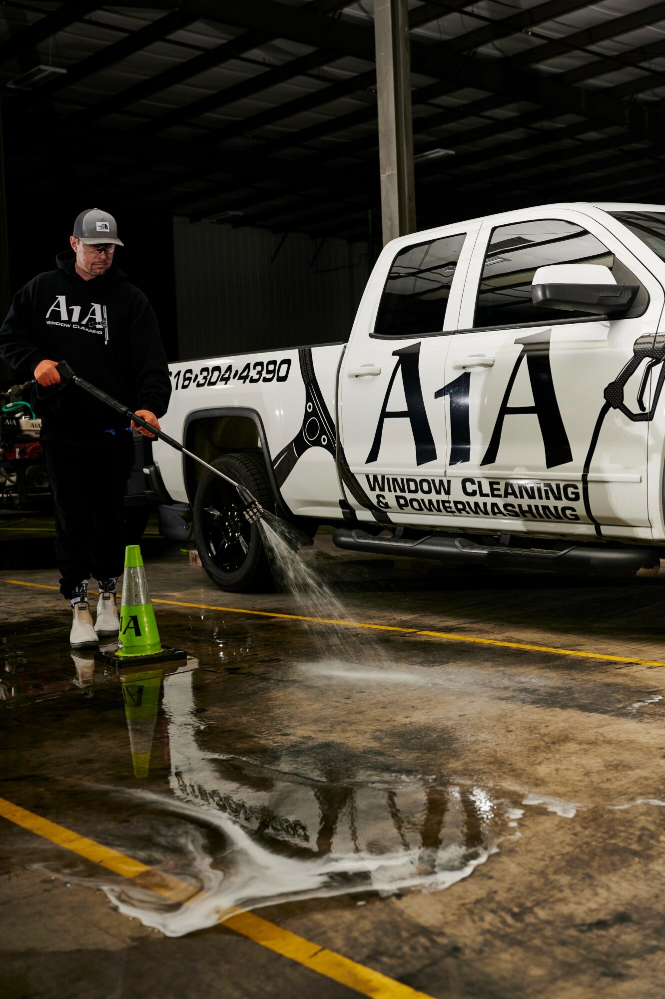 Person cleaning a white truck with a pressure washer inside a warehouse, water on the floor, and cleaning supplies nearby.