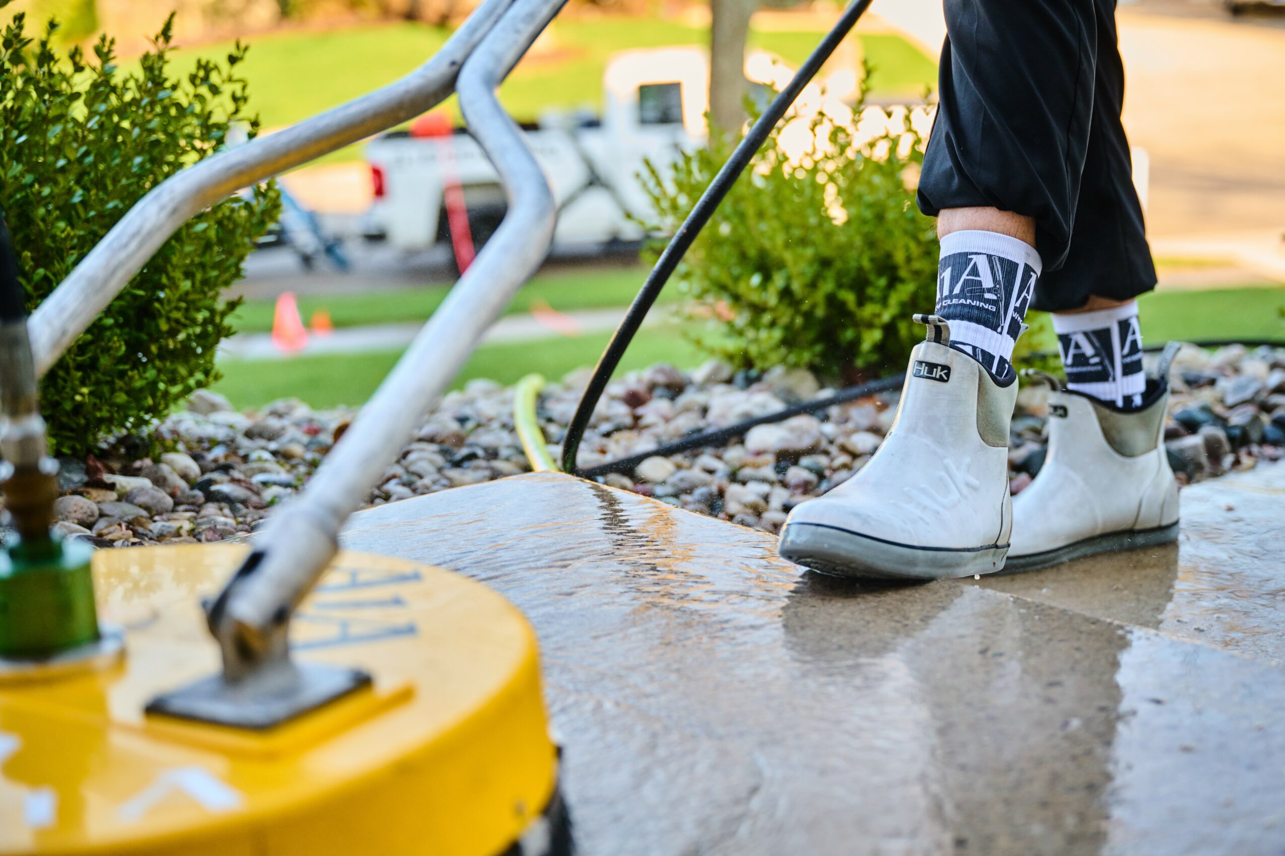 Person wearing boots and gloves using a sprayer on a wet sidewalk outdoors, with bushes and a vehicle in the background.