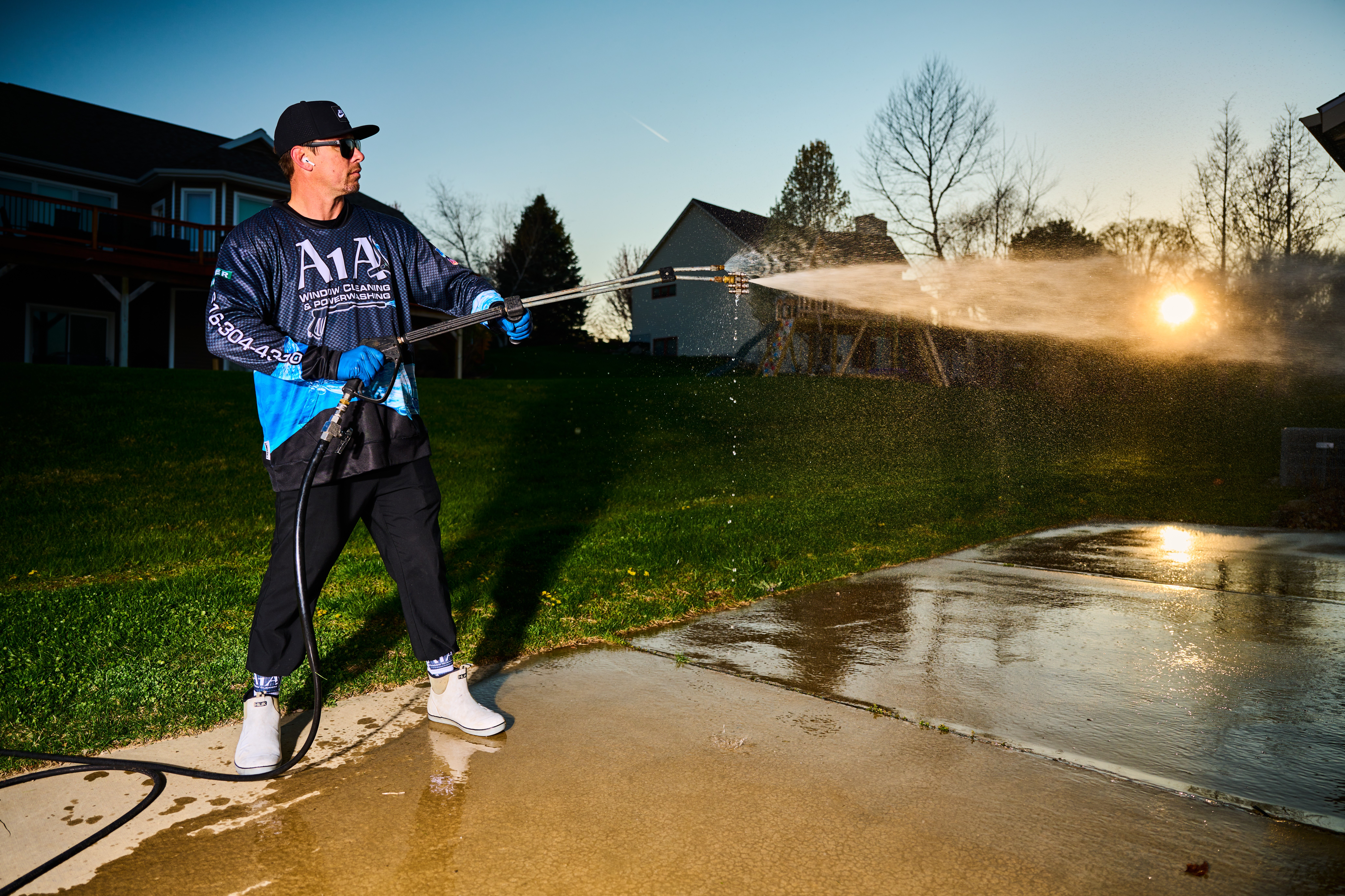 Person wearing a dark jacket and cap sprays water on a concrete surface outdoors during sunset.