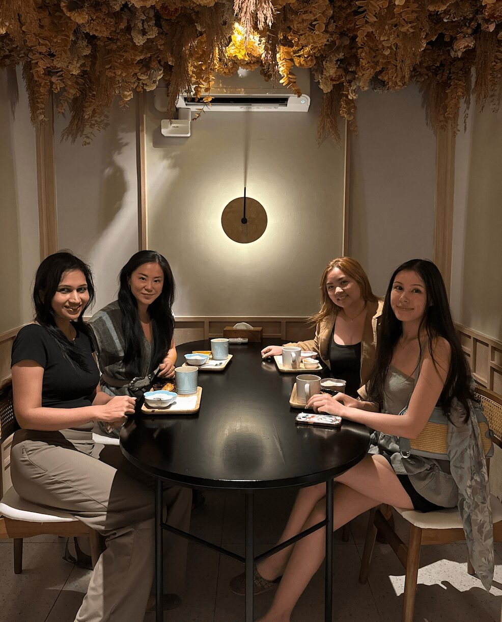 Four women sit around a black table in a room with a hanging light fixture and floral ceiling decor.