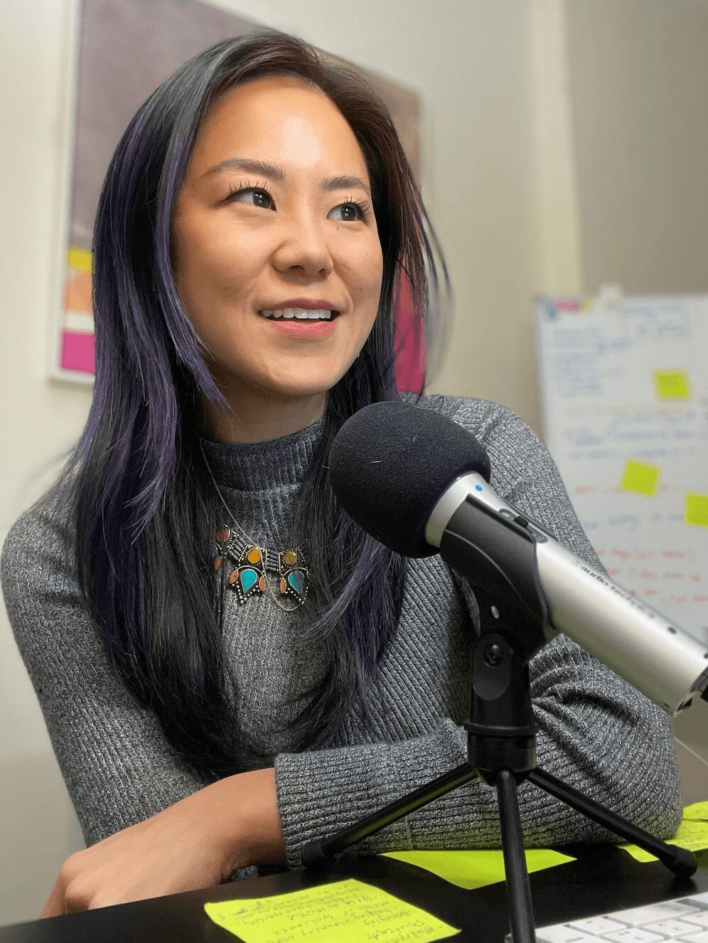 Woman with long dark hair speaking into a microphone during an interview or podcast, with a whiteboard and colorful sticky notes in the background.