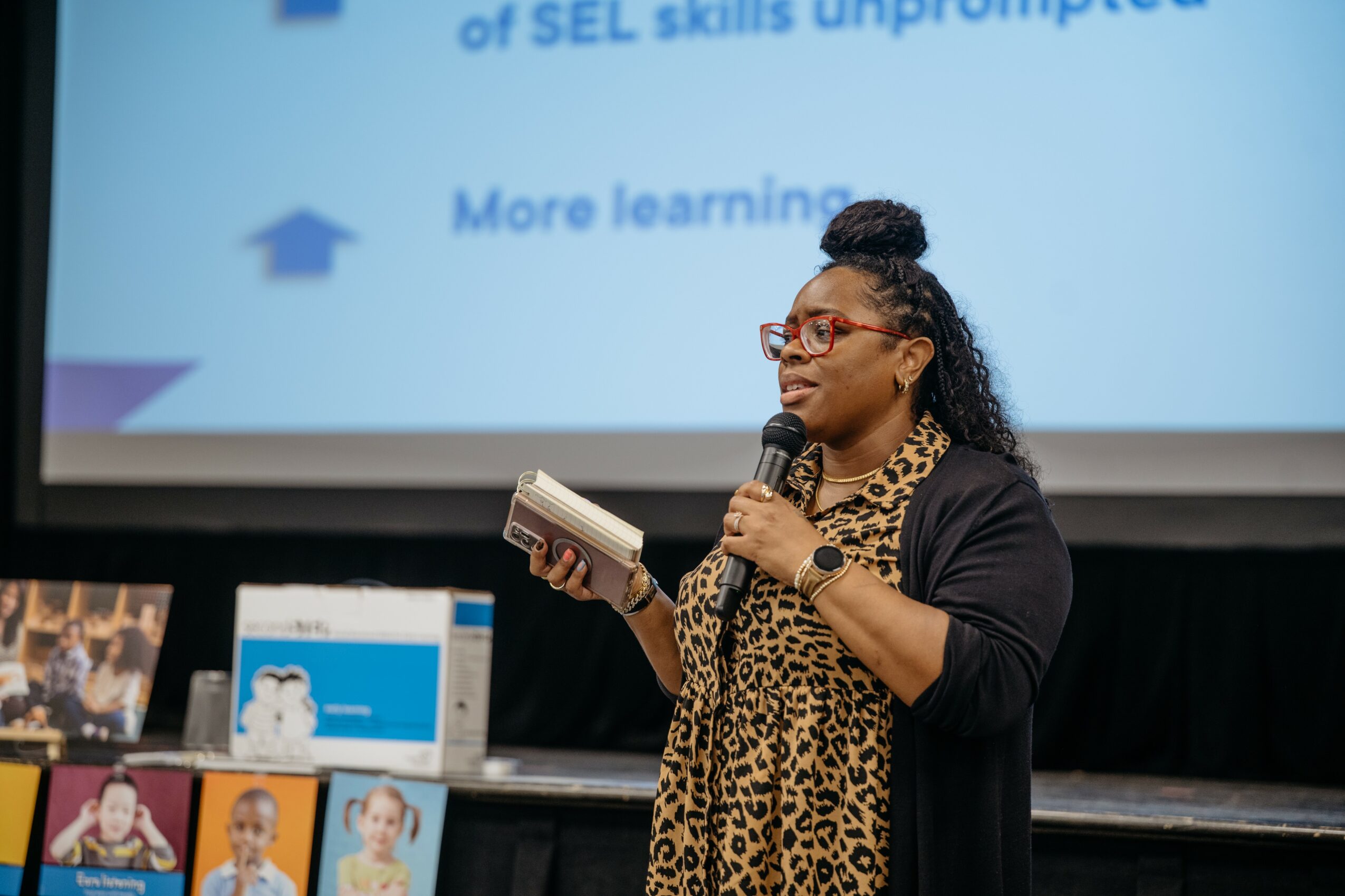 Woman speaking into microphone, holding a book, in front of a presentation screen, wearing glasses and a leopard print scarf.