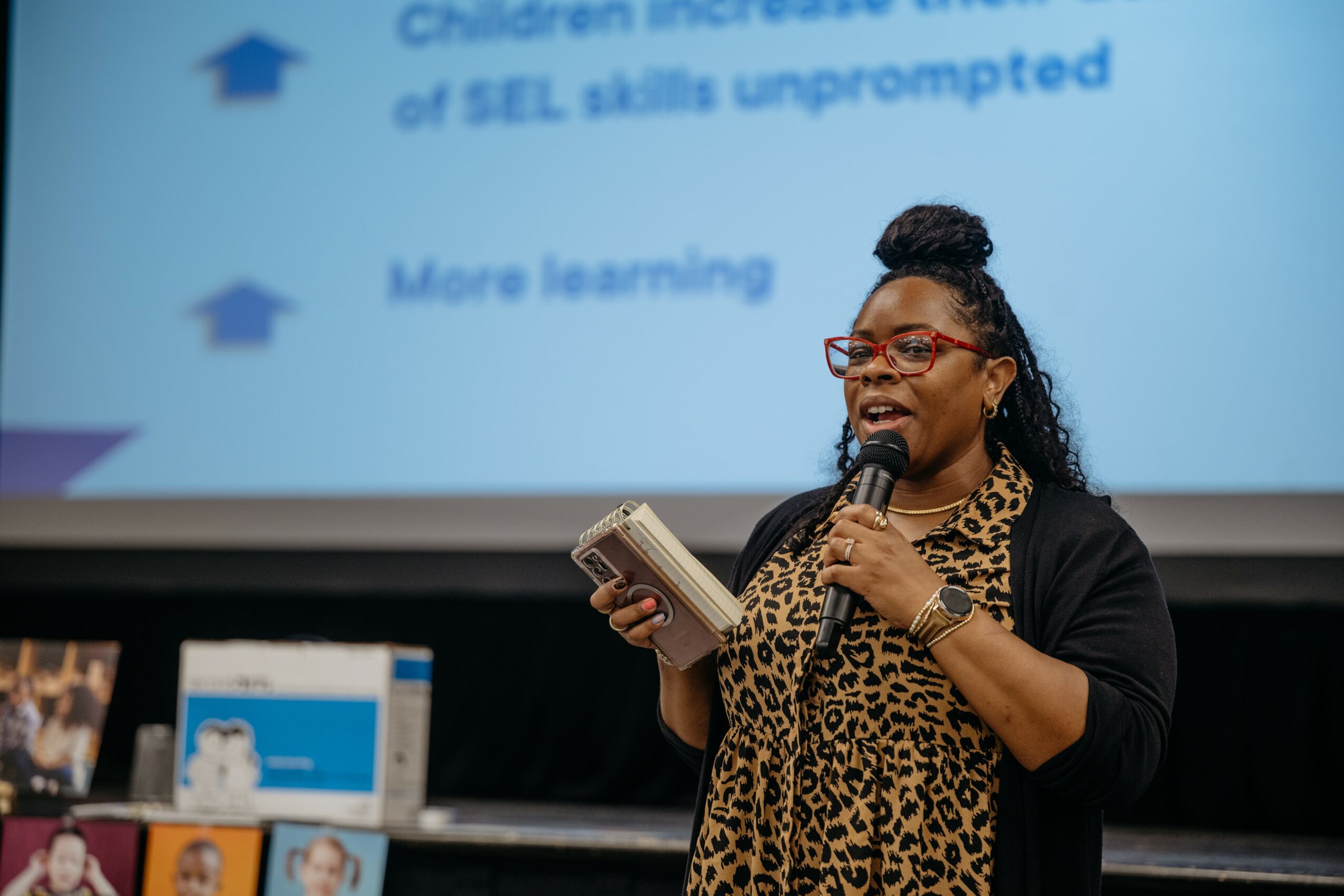 Woman speaking into microphone, holding notes, in front of a presentation screen with text, wearing glasses and a leopard print scarf.