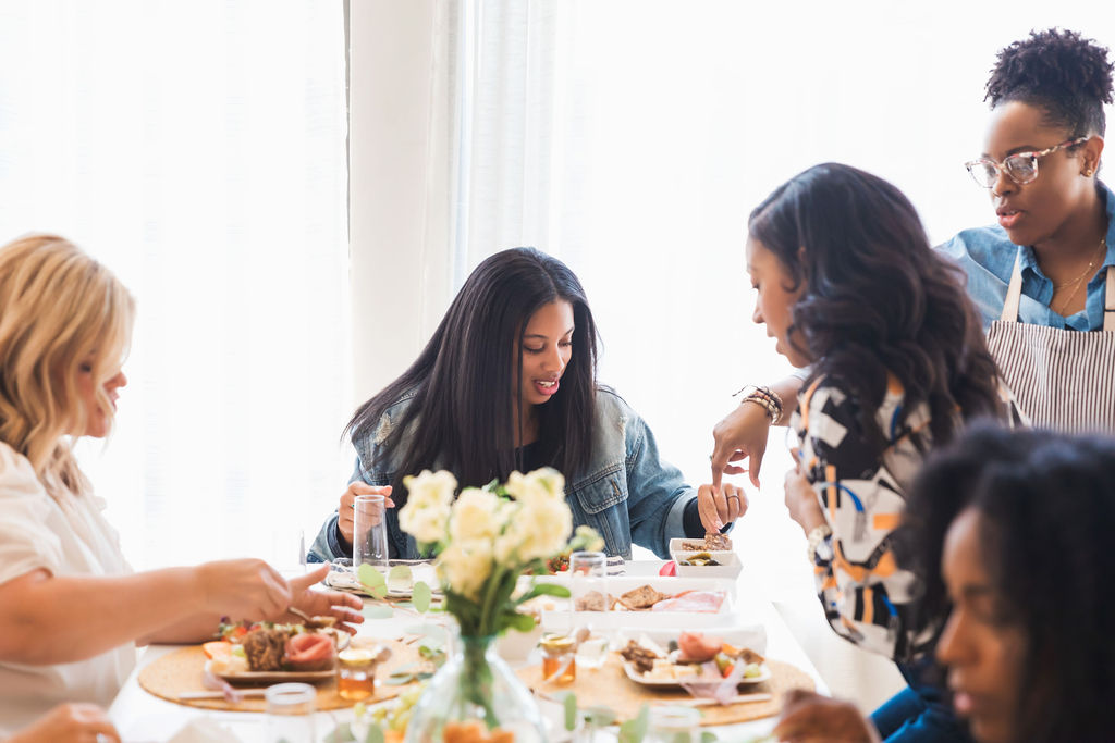 Group of women gathered around a table with food, a woman serving, and a vase with white flowers, in a bright room.
