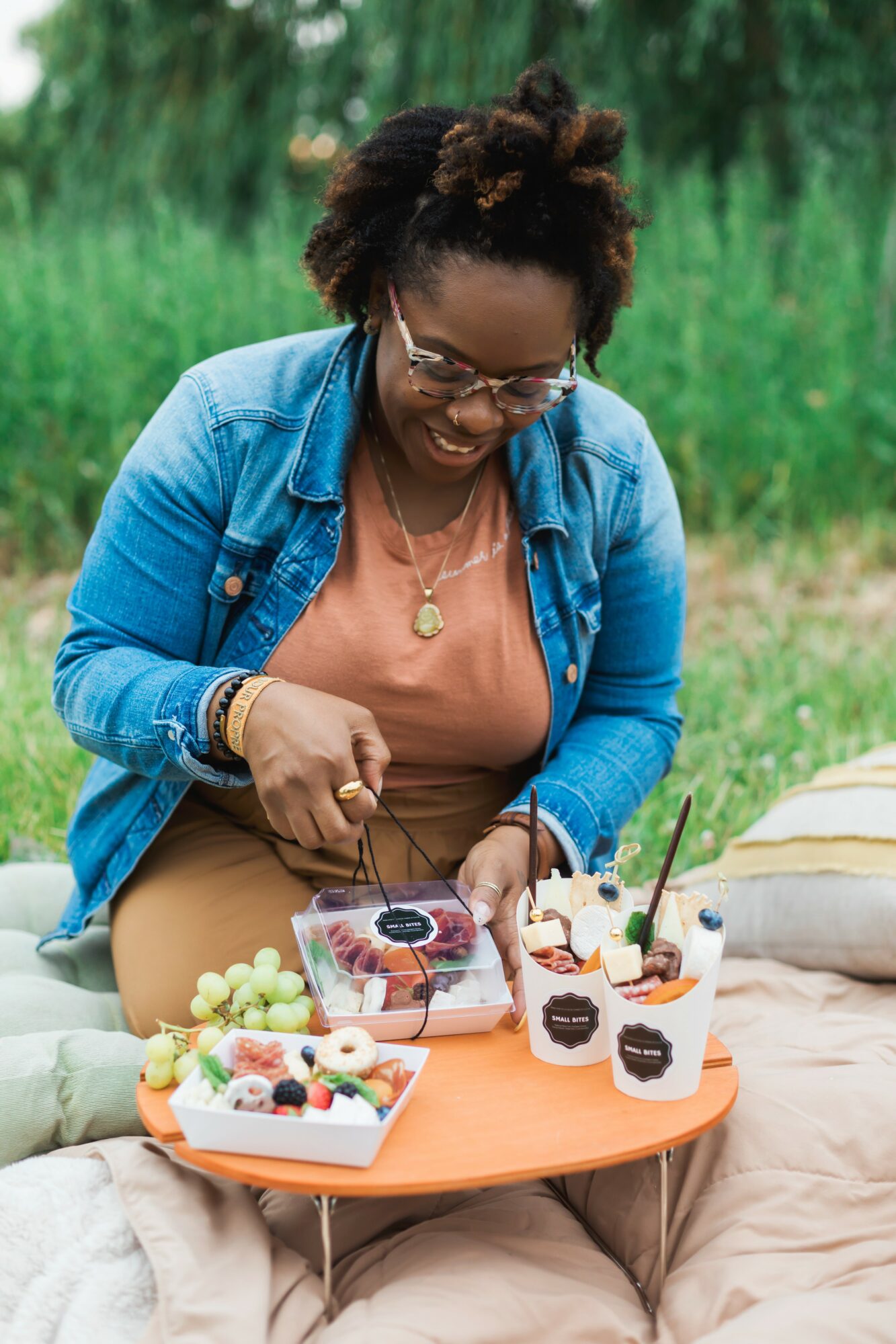 Woman with glasses and curly hair, wearing a denim jacket and orange top, smiling at a picnic table with desserts.