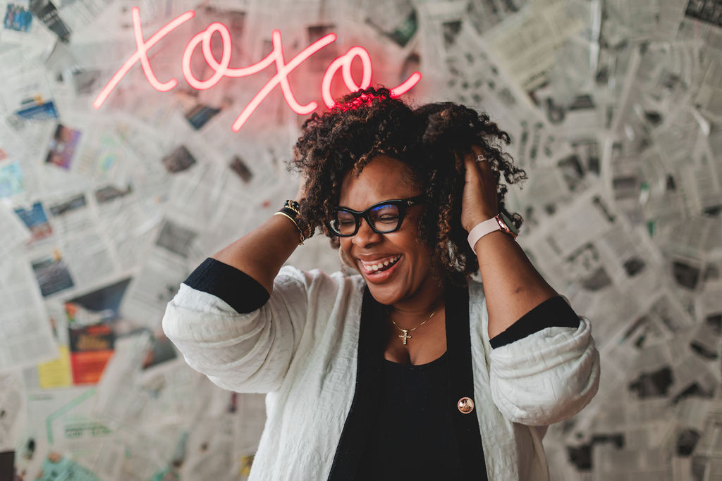 Woman with curly hair and glasses smiling, adjusting her hair, in front of a newspaper-covered wall with pink neon text.