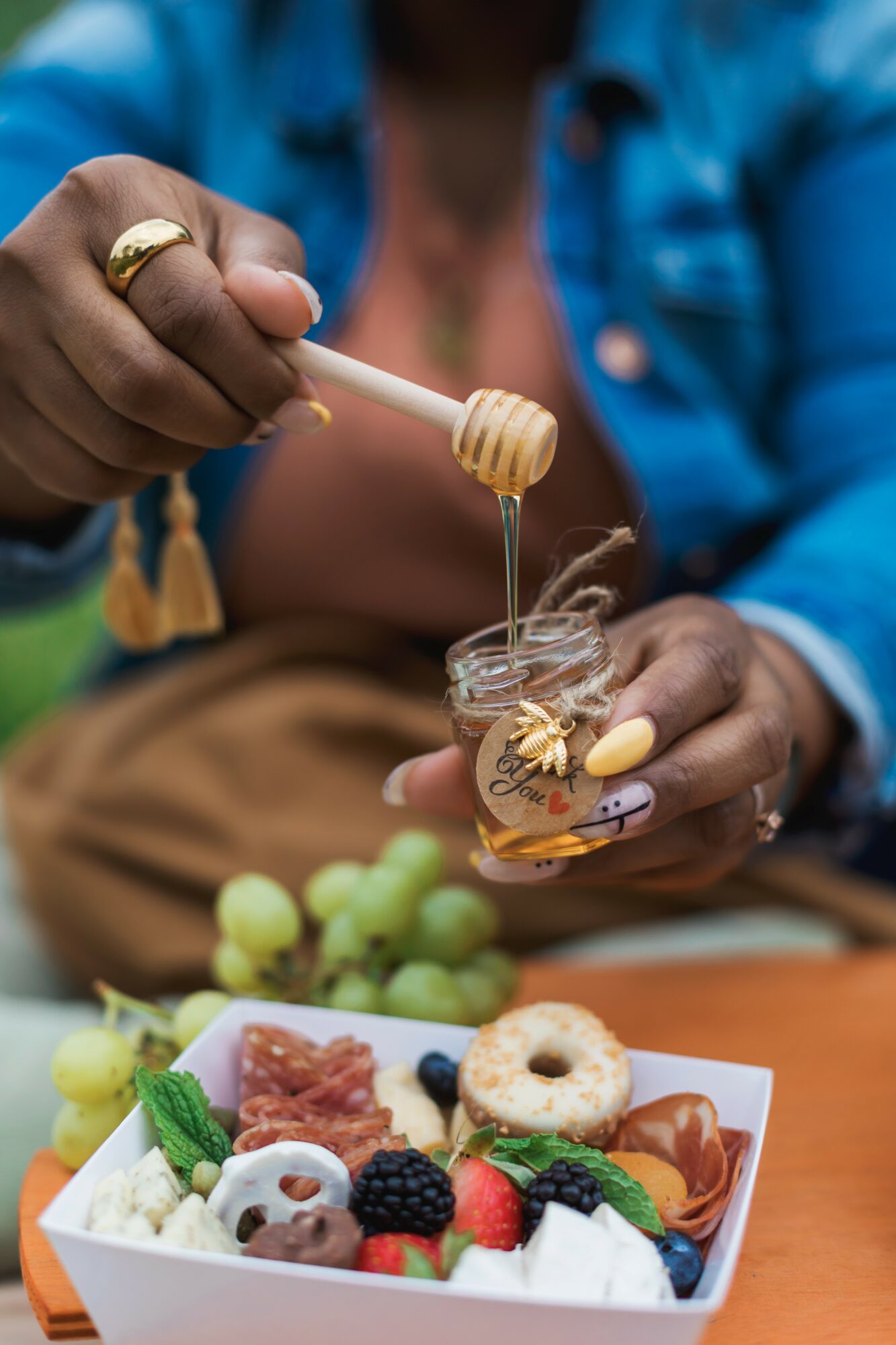 Person holding a honey dipper over a jar of honey, with a plate of assorted desserts and grapes nearby.