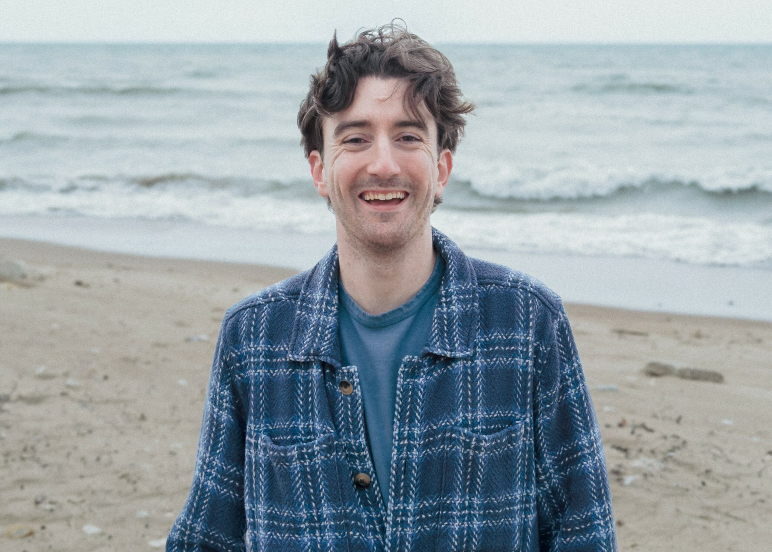 Young man smiling on a beach with ocean waves in the background.