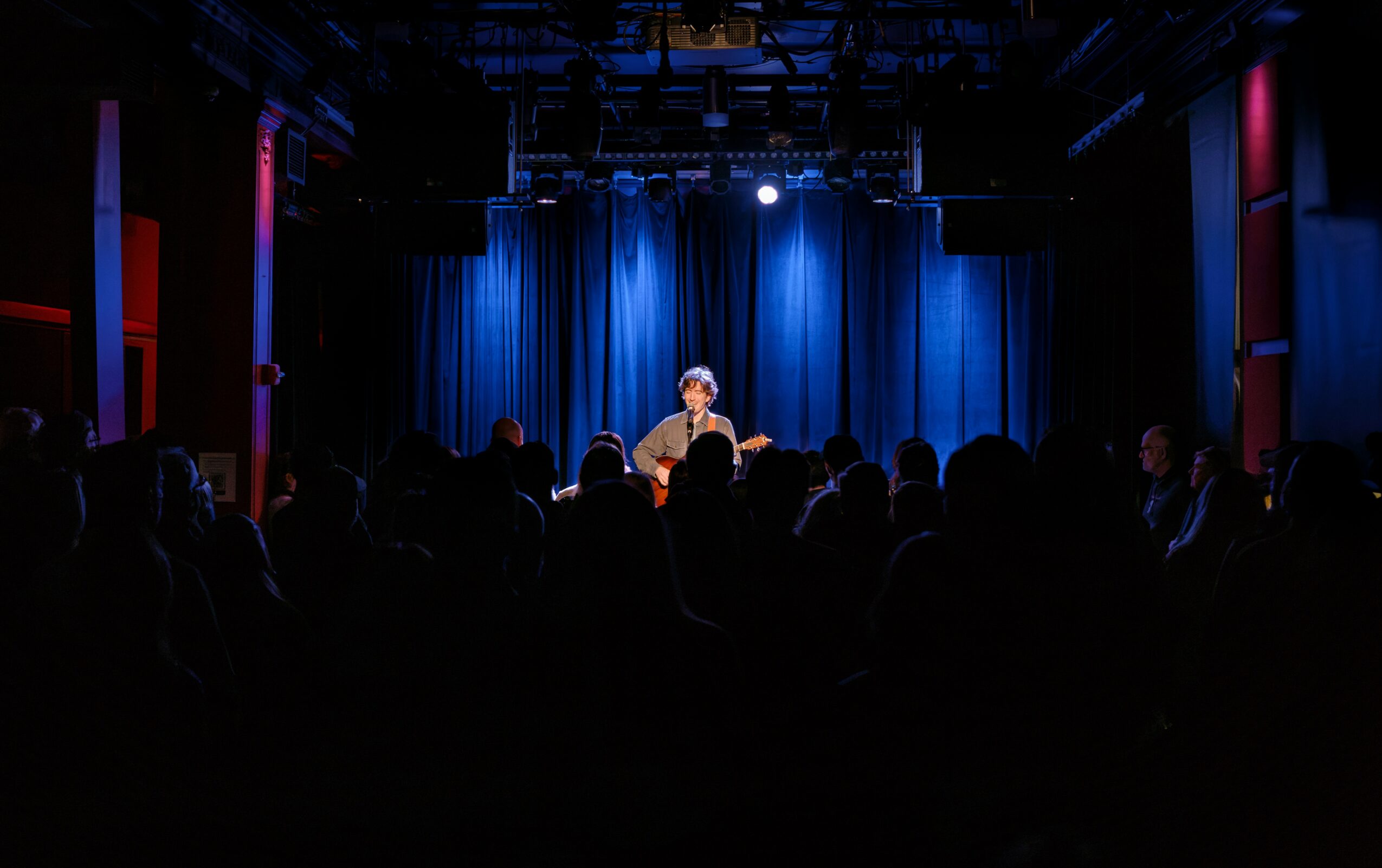 Performer on stage with blue curtains, audience in foreground, dim lighting, theater setting.