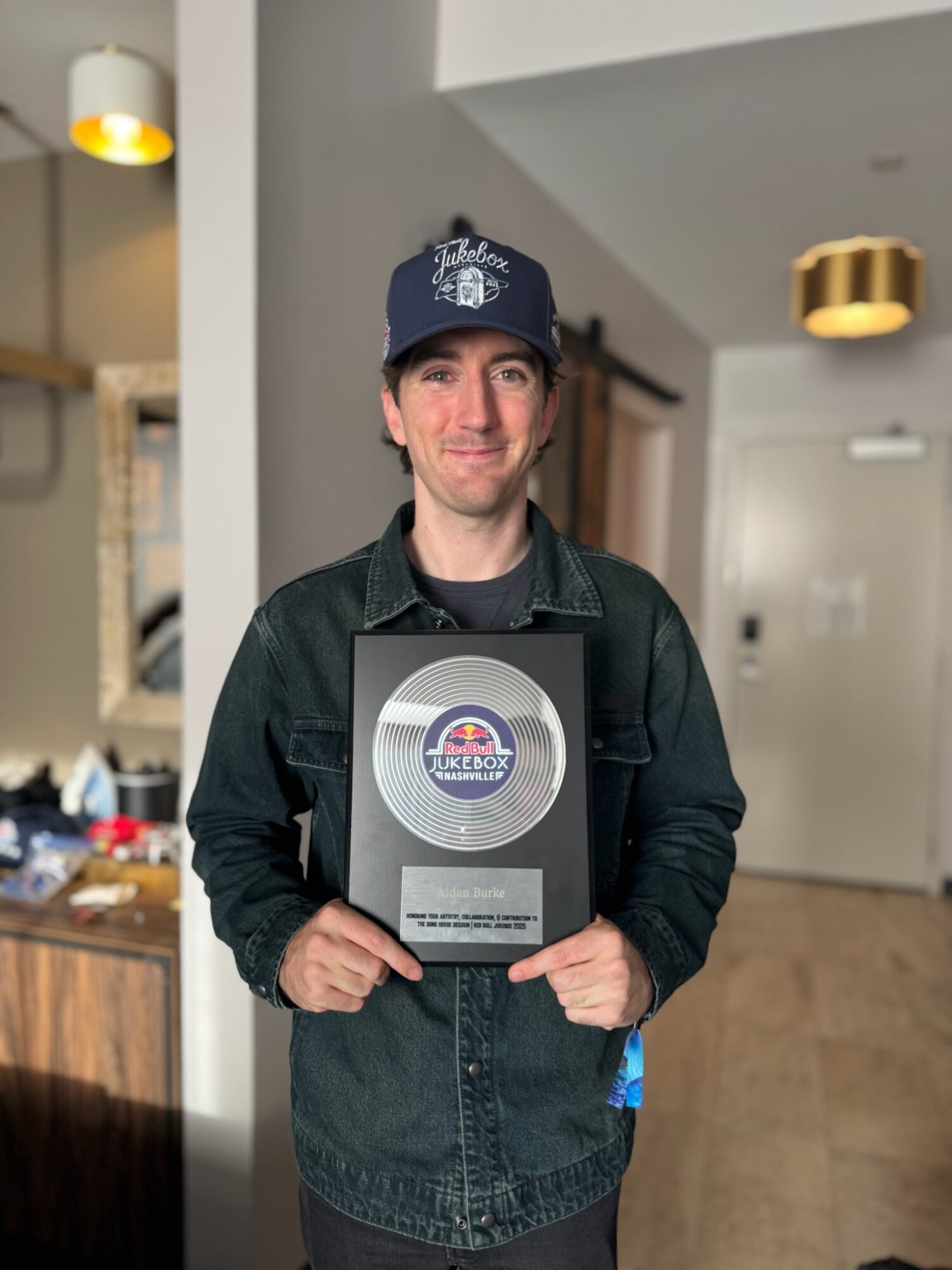 Young man holding a plaque, wearing a cap and denim jacket, standing indoors with modern decor.