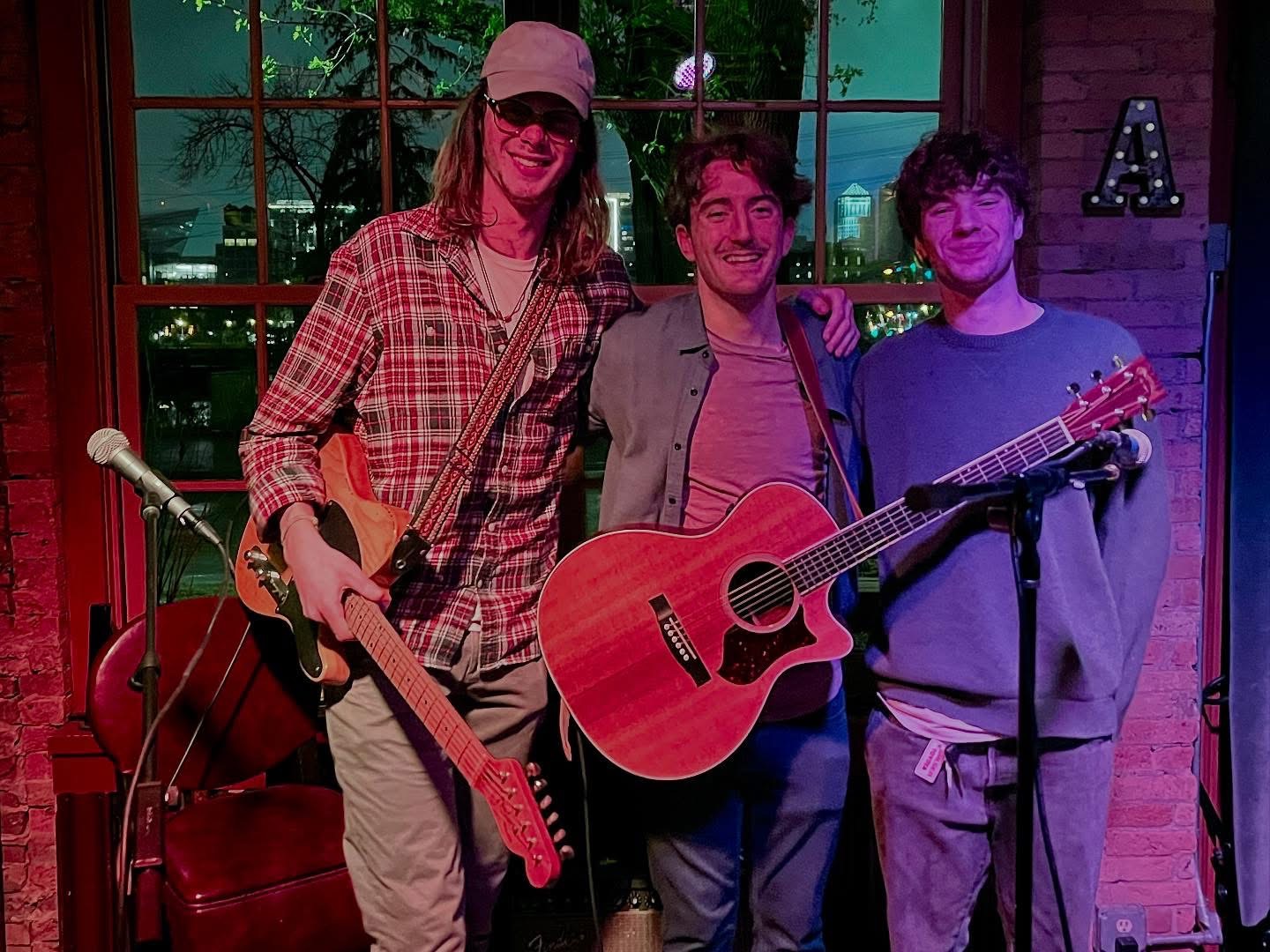 Three people holding guitars and microphone stand in a room with large window and brick wall.