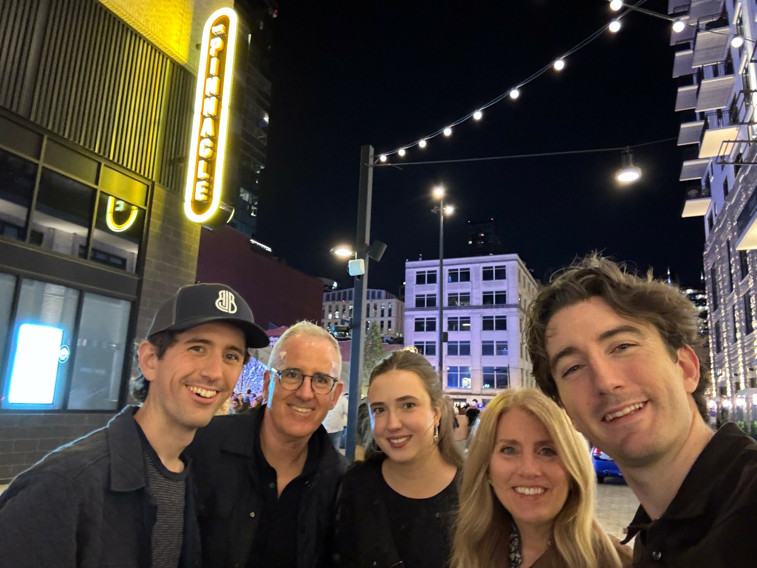 Group of five people smiling outdoors at night with city buildings and string lights in background.
