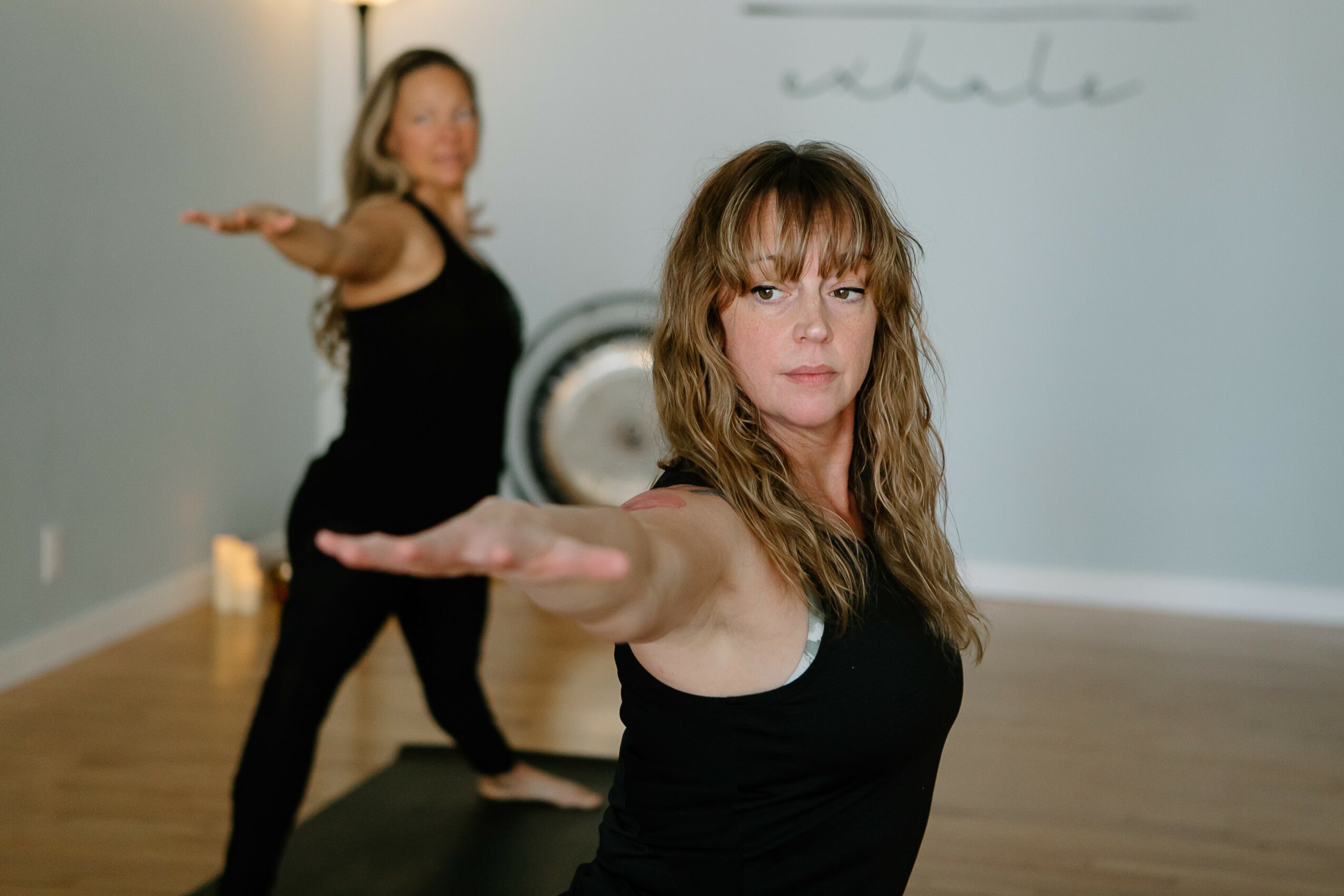 Two women practicing yoga in a studio, one in the foreground and one in the background, both in black workout clothes.