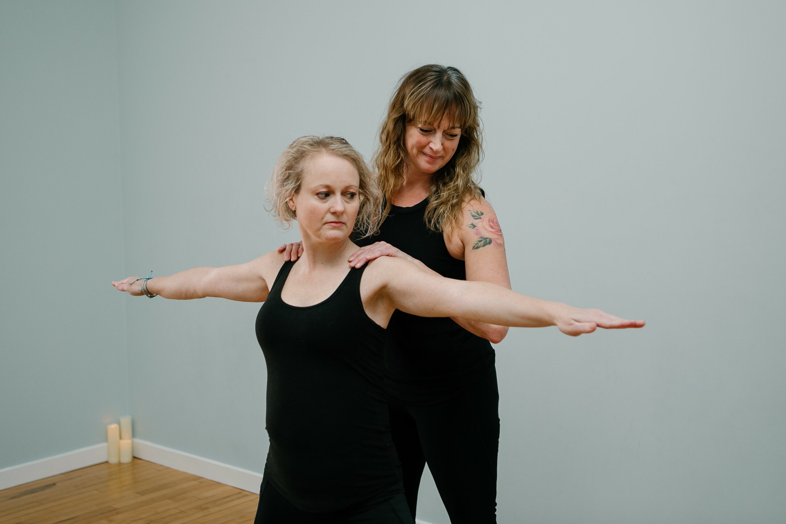 Two women in black clothing stand with arms extended sideways in a room with light-colored walls and wooden floor.