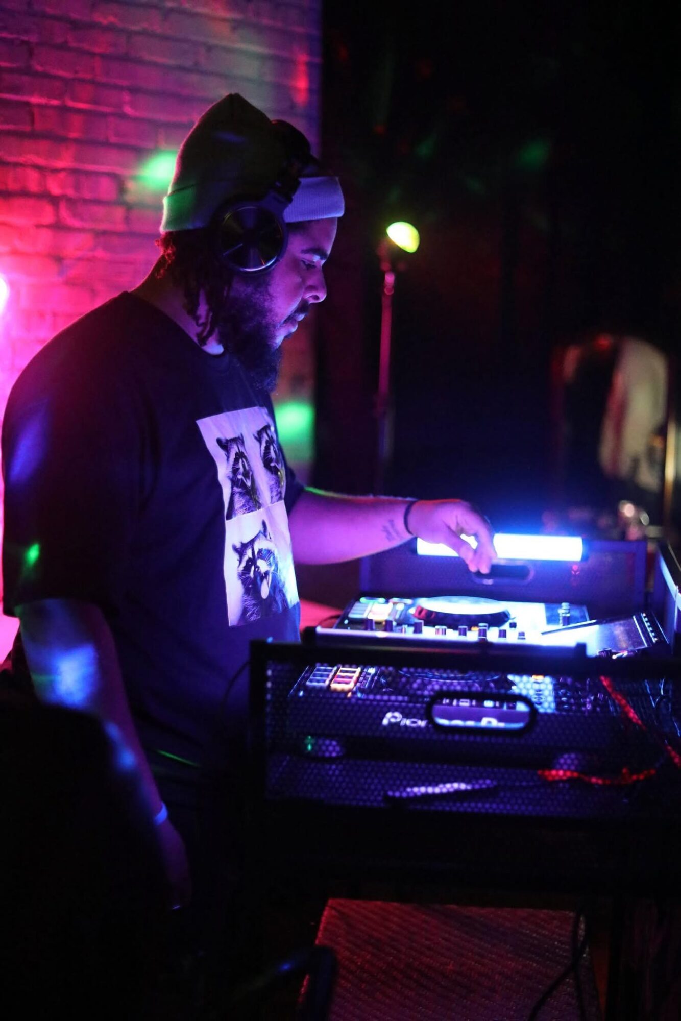 Person with headphones and beard operating DJ equipment in a dimly lit venue with colorful lights.