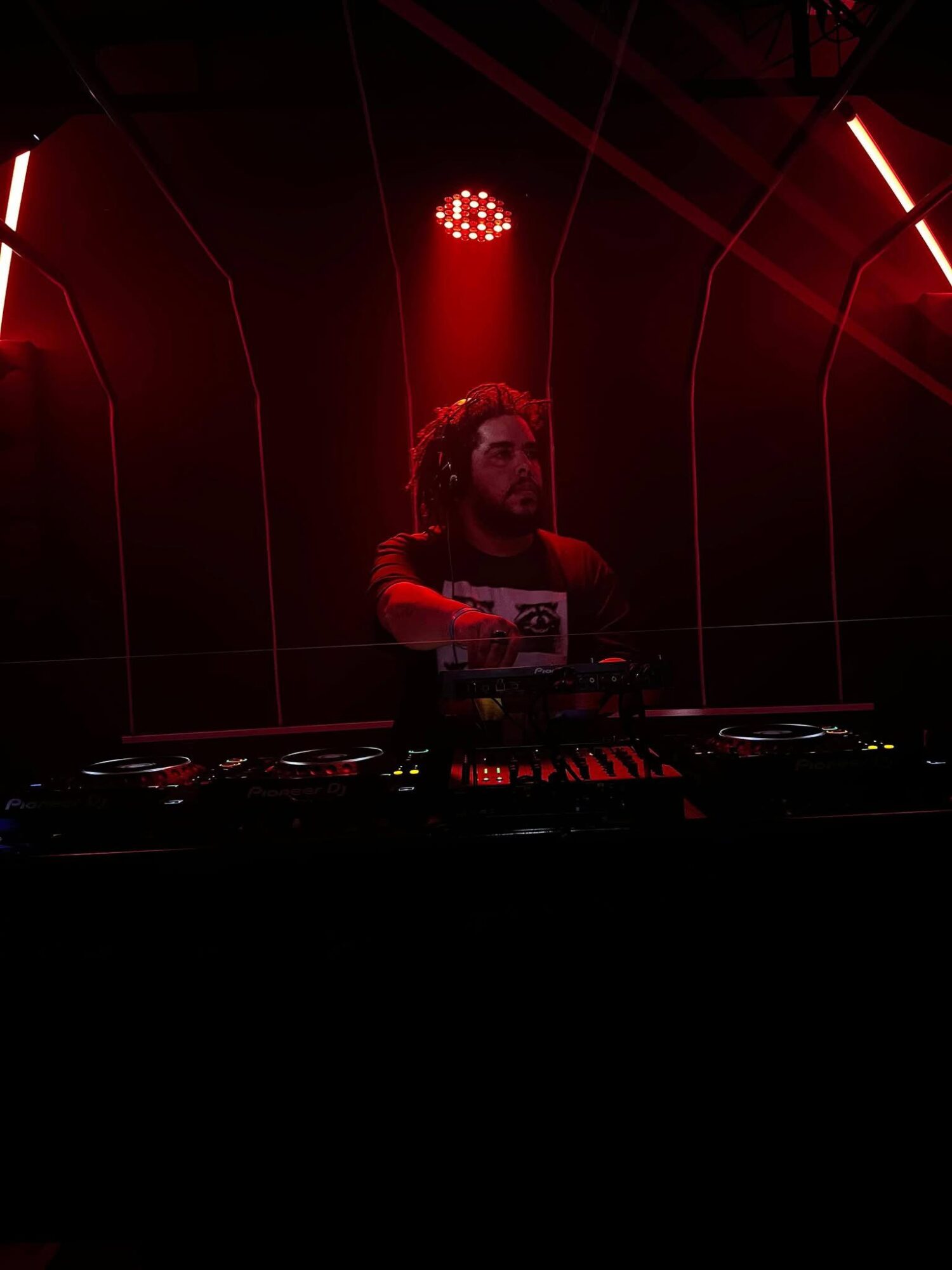 DJ with curly hair and beard performing in a dark, red-lit environment with equipment in front.