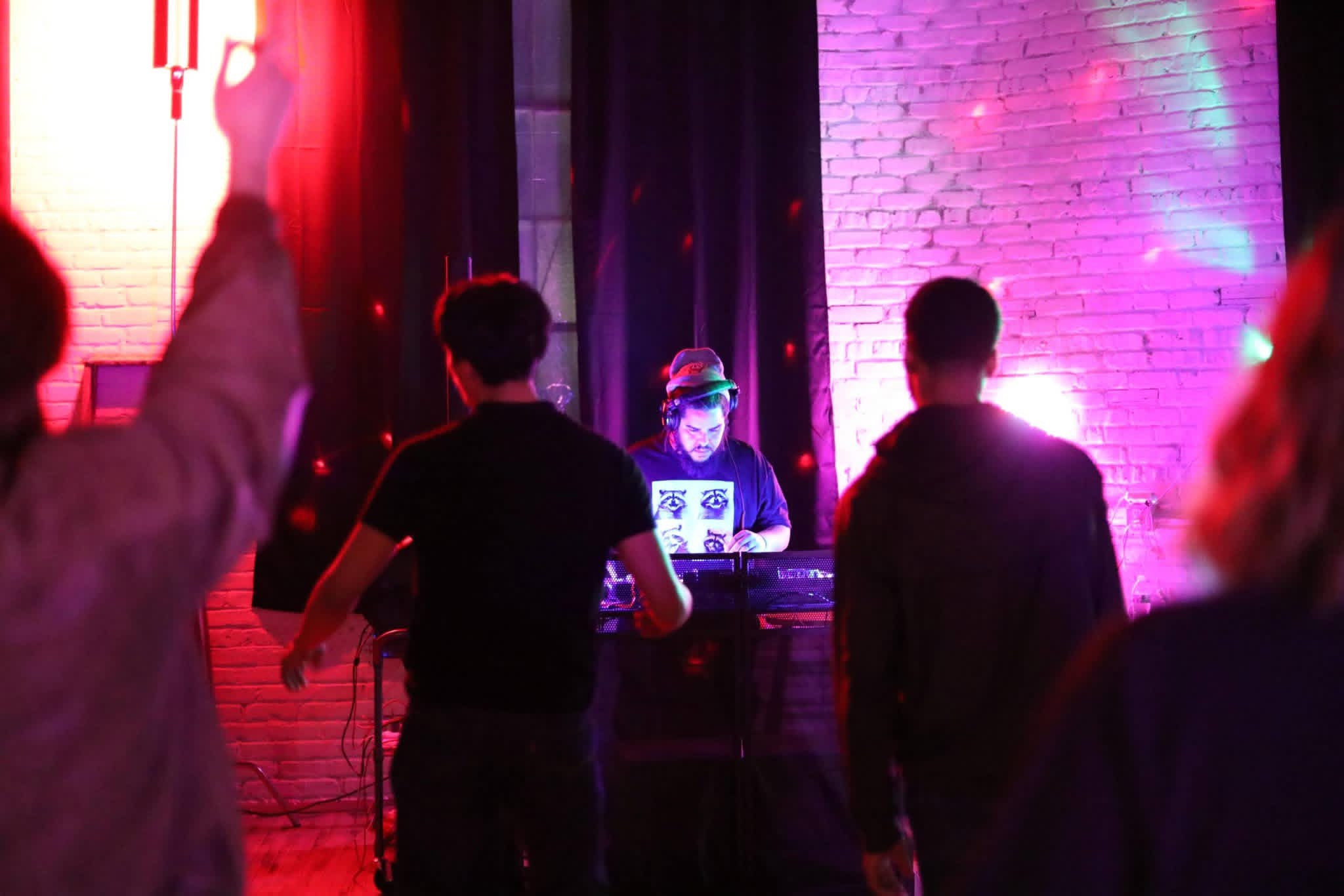 Person DJing on stage with colorful lighting, audience watching, brick wall background, dark setting, lively atmosphere.