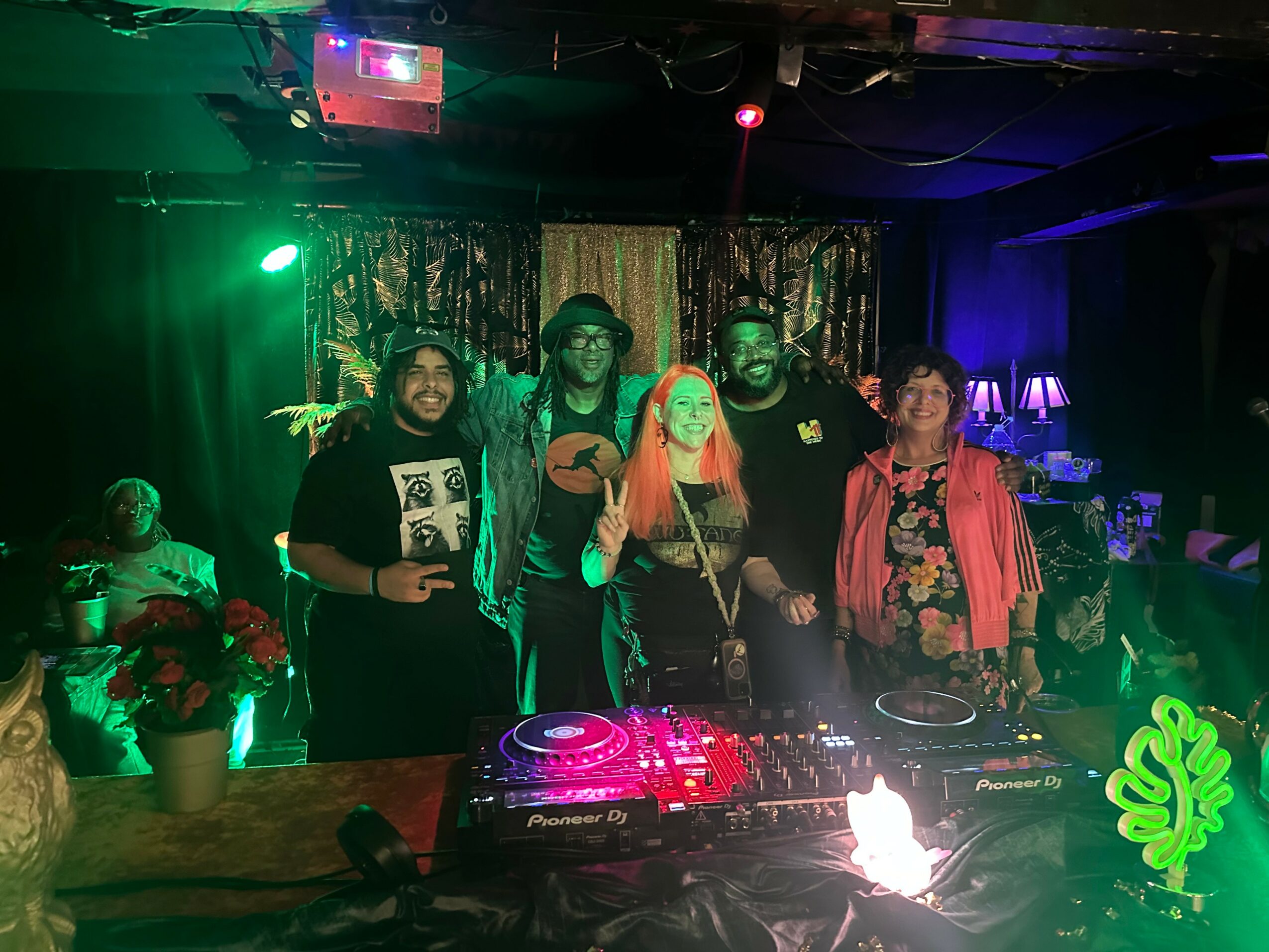 Group of five people standing behind DJ equipment in a dimly lit club with colorful lights, smiling and posing for the photo.