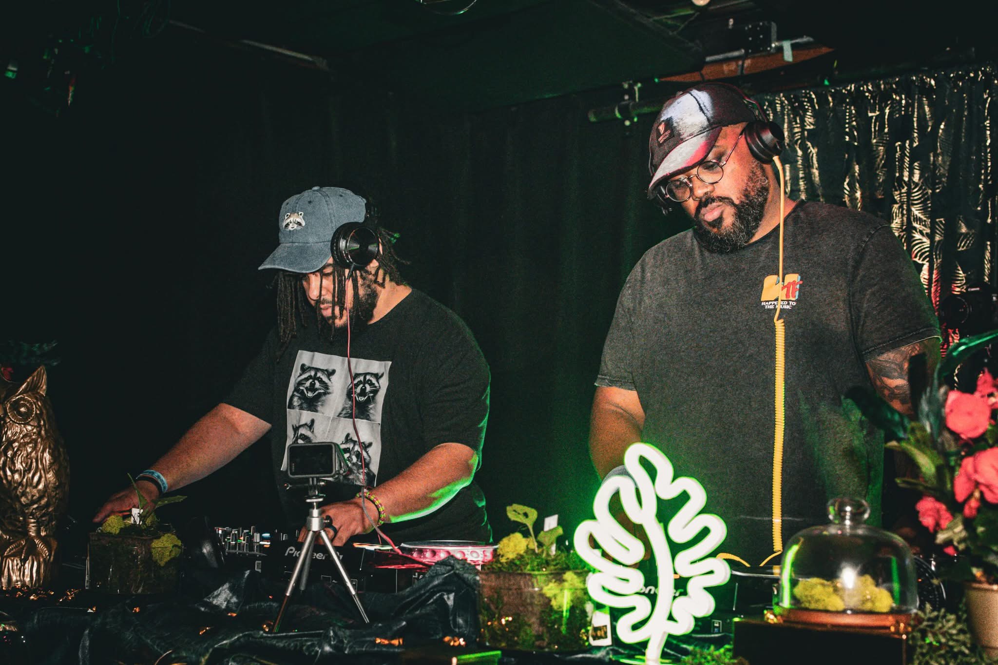 Two DJs perform behind a mixing console with plants and decorative items in front, dark background with curtains, one wearing a cap.