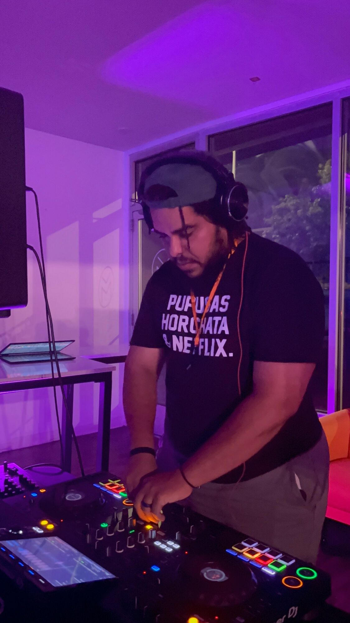 Person DJing with headphones at a mixing console, purple lighting, large window in background