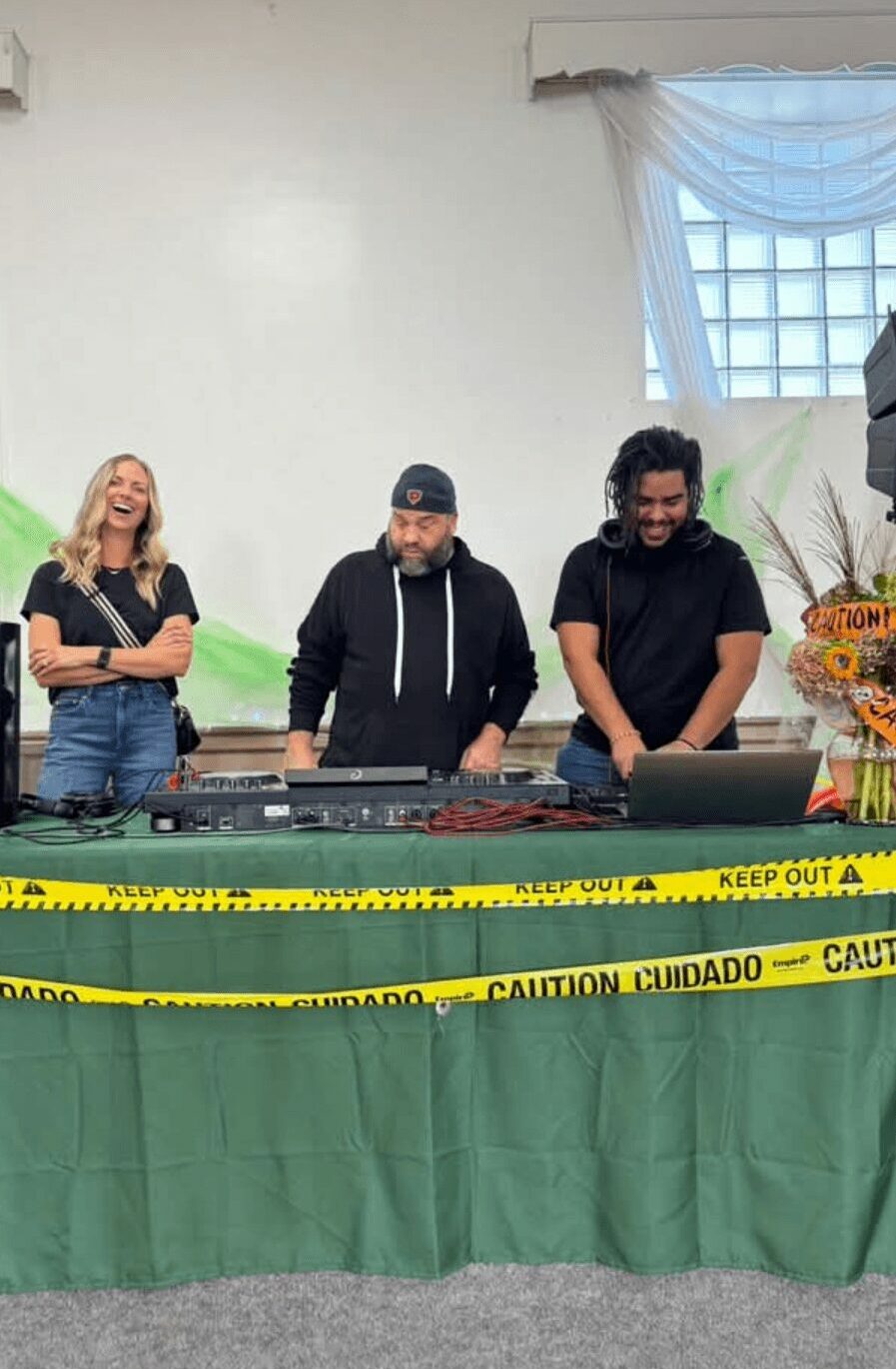 Three people standing behind a table with yellow caution tape, a woman on the left, two men in the middle and right, in a room with a window and curtain.