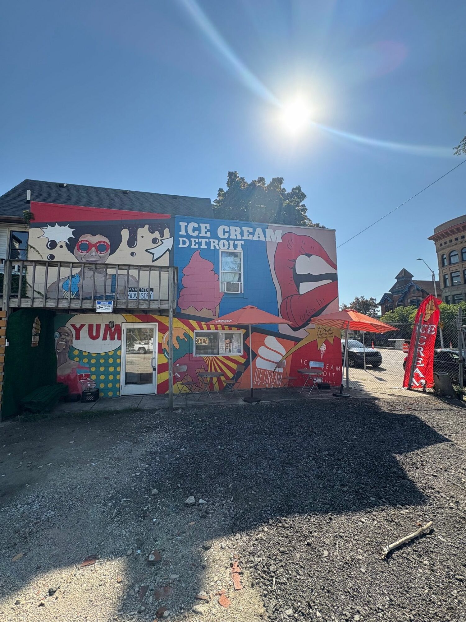 Ice cream stand with colorful artwork, a red umbrella, and a sign reading 'YUM' on a sunny day.