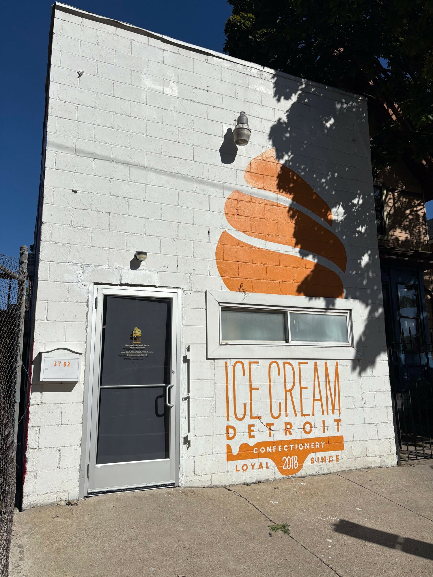 White brick wall with ice cream cone mural, window, and door, advertising ice cream flavors and prices, sidewalk in front.