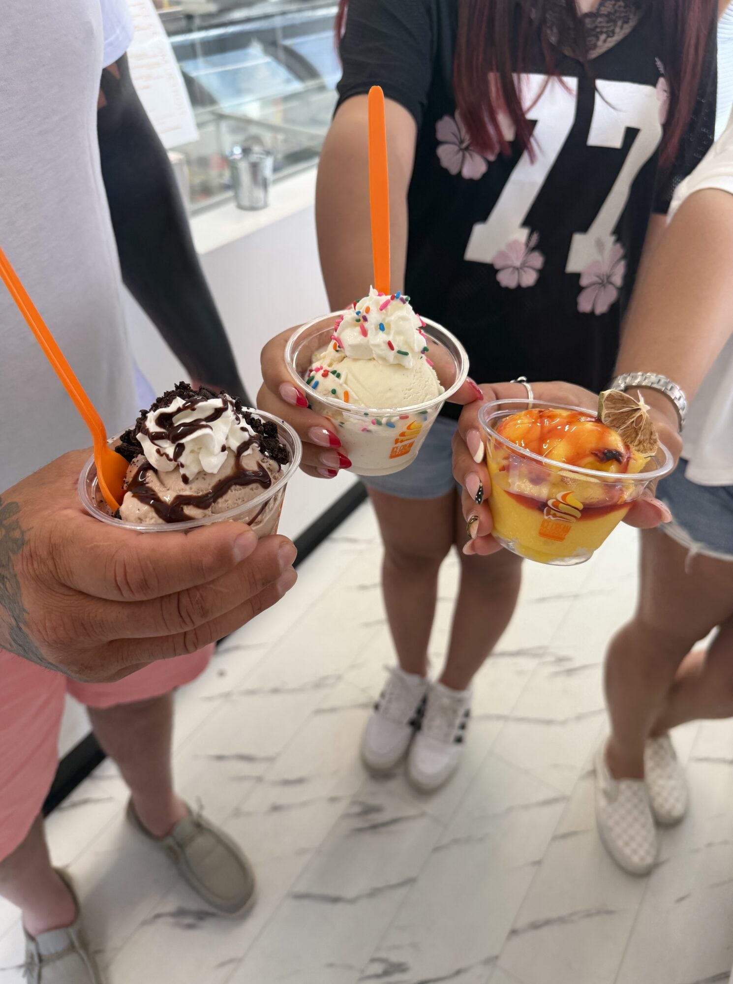 Three people holding cups of ice cream with toppings, standing on a white marble floor indoors.