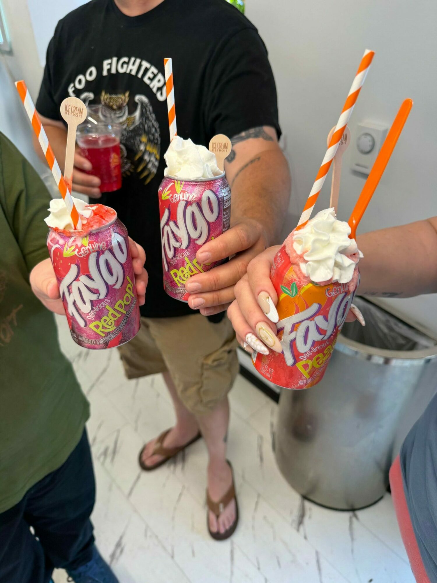 Two people hold colorful drinks with whipped cream and straws, standing on a tiled floor, with one person wearing flip-flops.