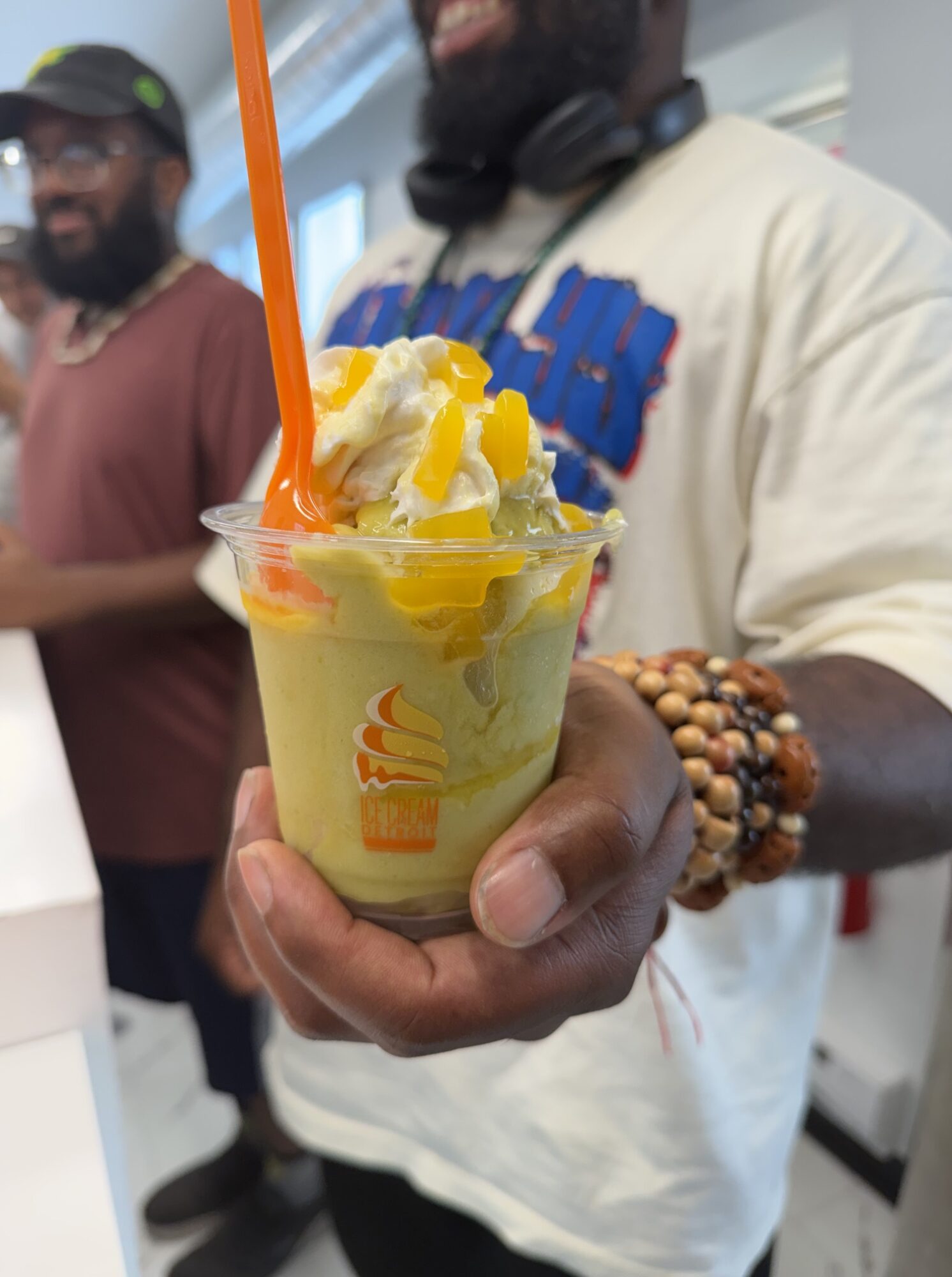 Person holding a yellow ice cream with a straw, with another person in the background, indoors.