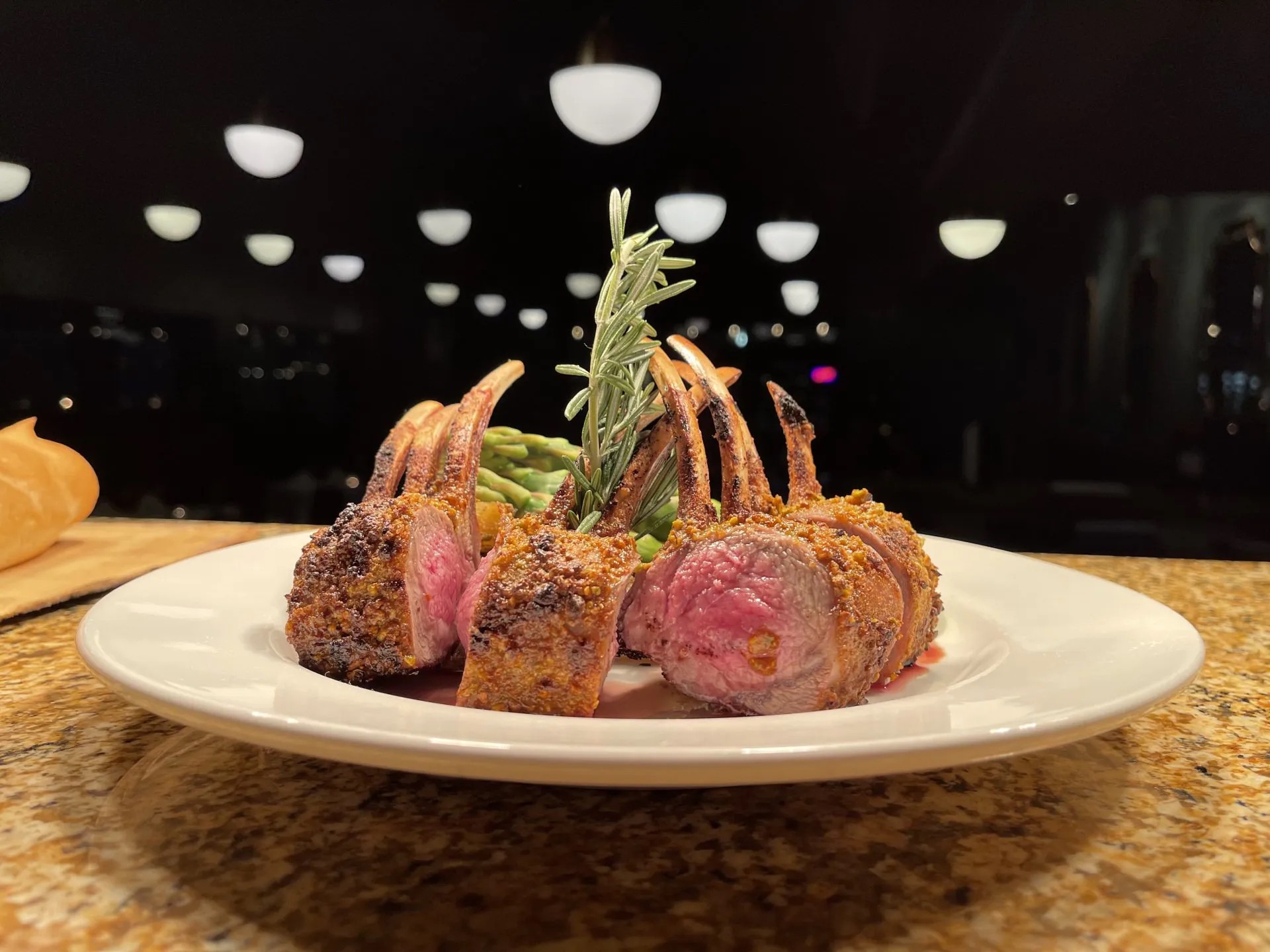 Plate with cooked meat, garnished with a sprig of herbs, on a table with a dark background and ceiling lights.