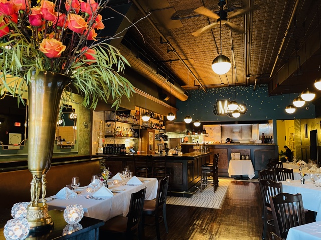 Elegant restaurant interior with tables set for dining, a large floral arrangement, and warm lighting.