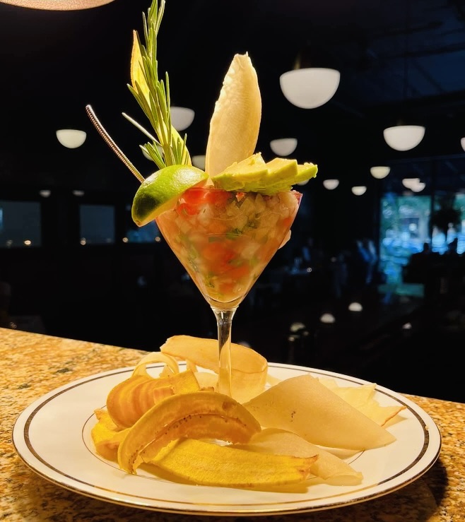 Colorful cocktail garnished with lime, pineapple, and herbs, served with sliced fruit on a white plate, in a dimly lit setting.