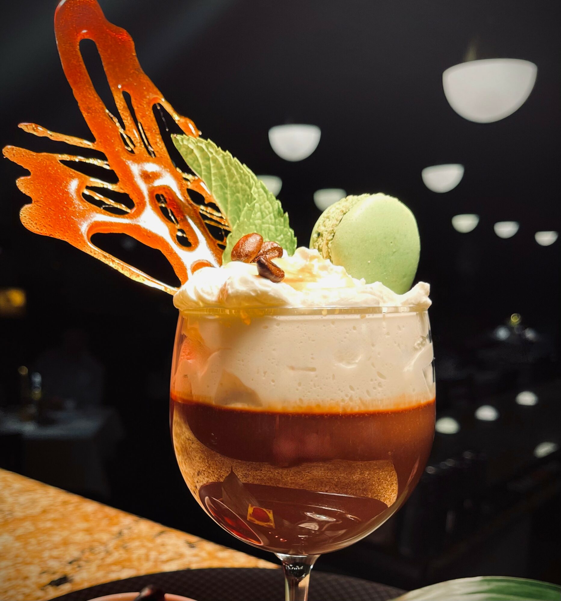 A dessert drink with whipped cream, a green macaron, and decorative caramel on a dark background.