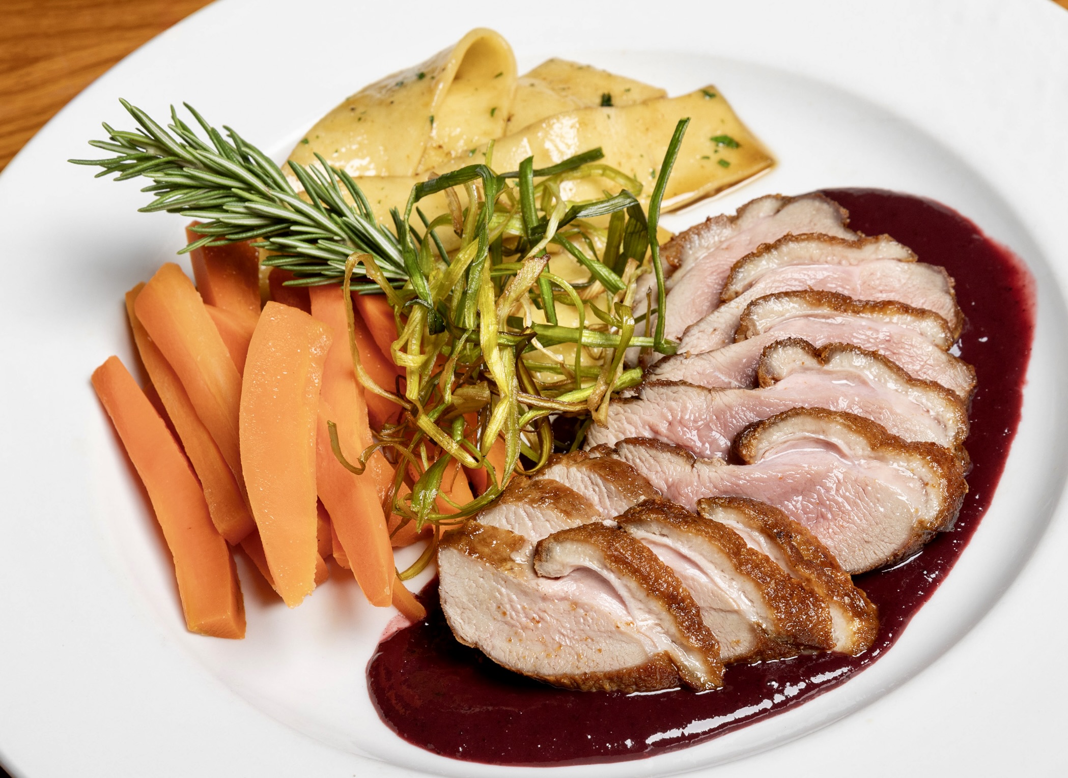 Sliced cooked meat with sauce, carrots, pasta, and greens on a white plate.