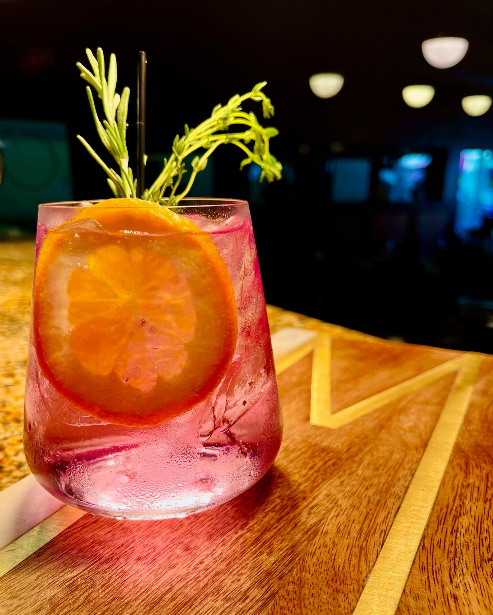 Pink glass with orange slice and green garnish on wooden surface, dark background with ceiling lights.
