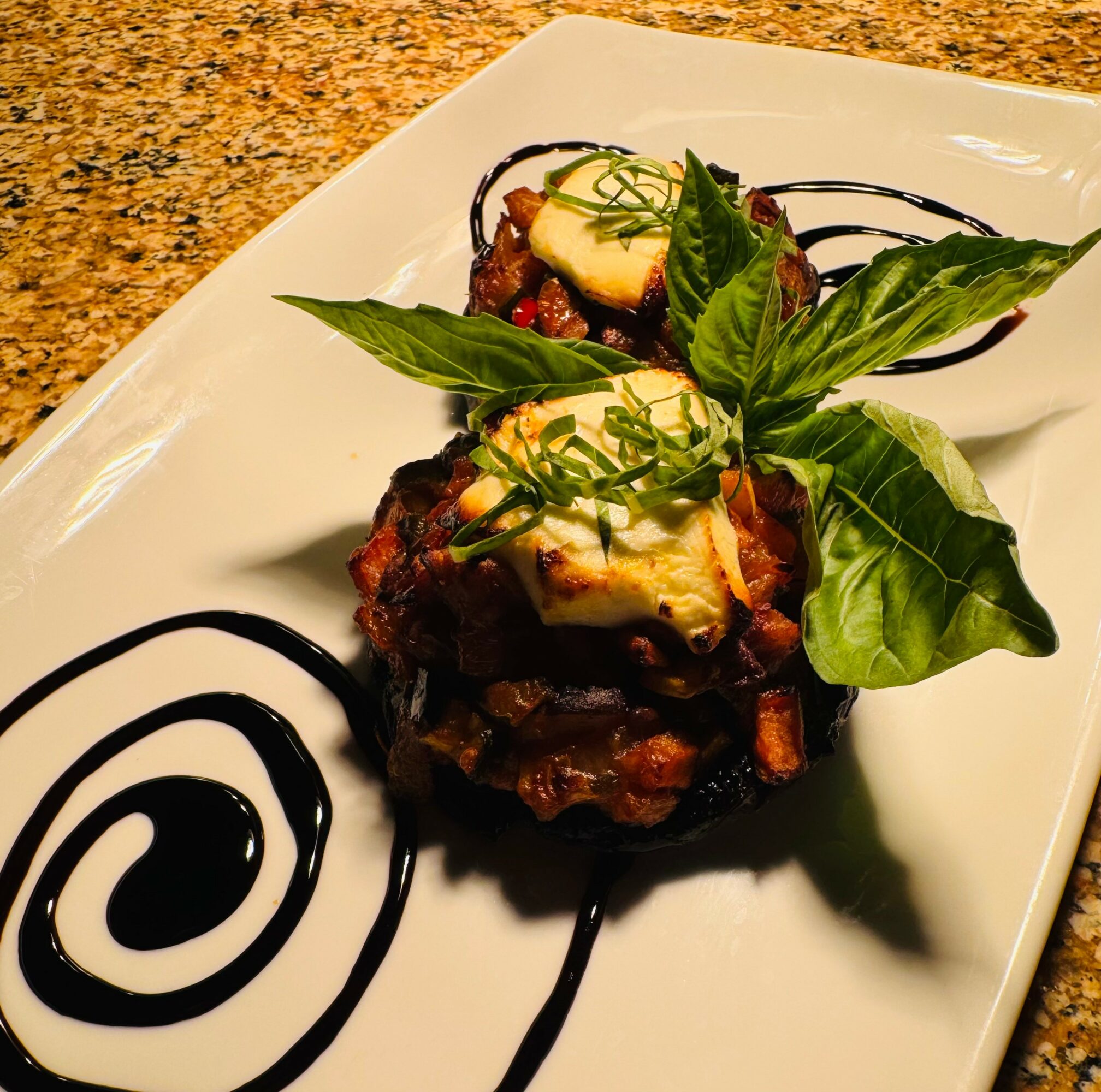 Plate with two pieces of food topped with cheese and herbs, garnished with basil leaves, and decorated with black sauce swirls.