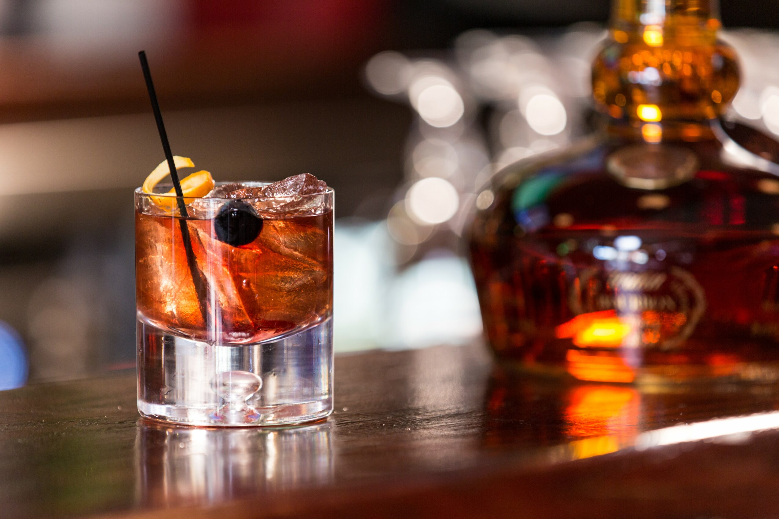 A cocktail glass with ice, a lemon slice, and a cherry, on a bar counter with a blurred background.