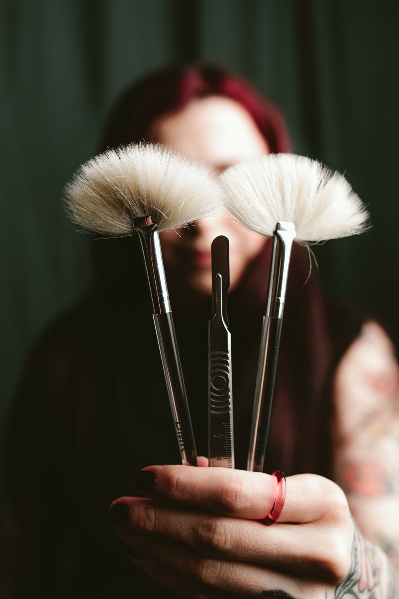 Person holding three makeup brushes with fluffy white bristles, blurred background, dark green curtain, hand with red ring, and tattoos visible.