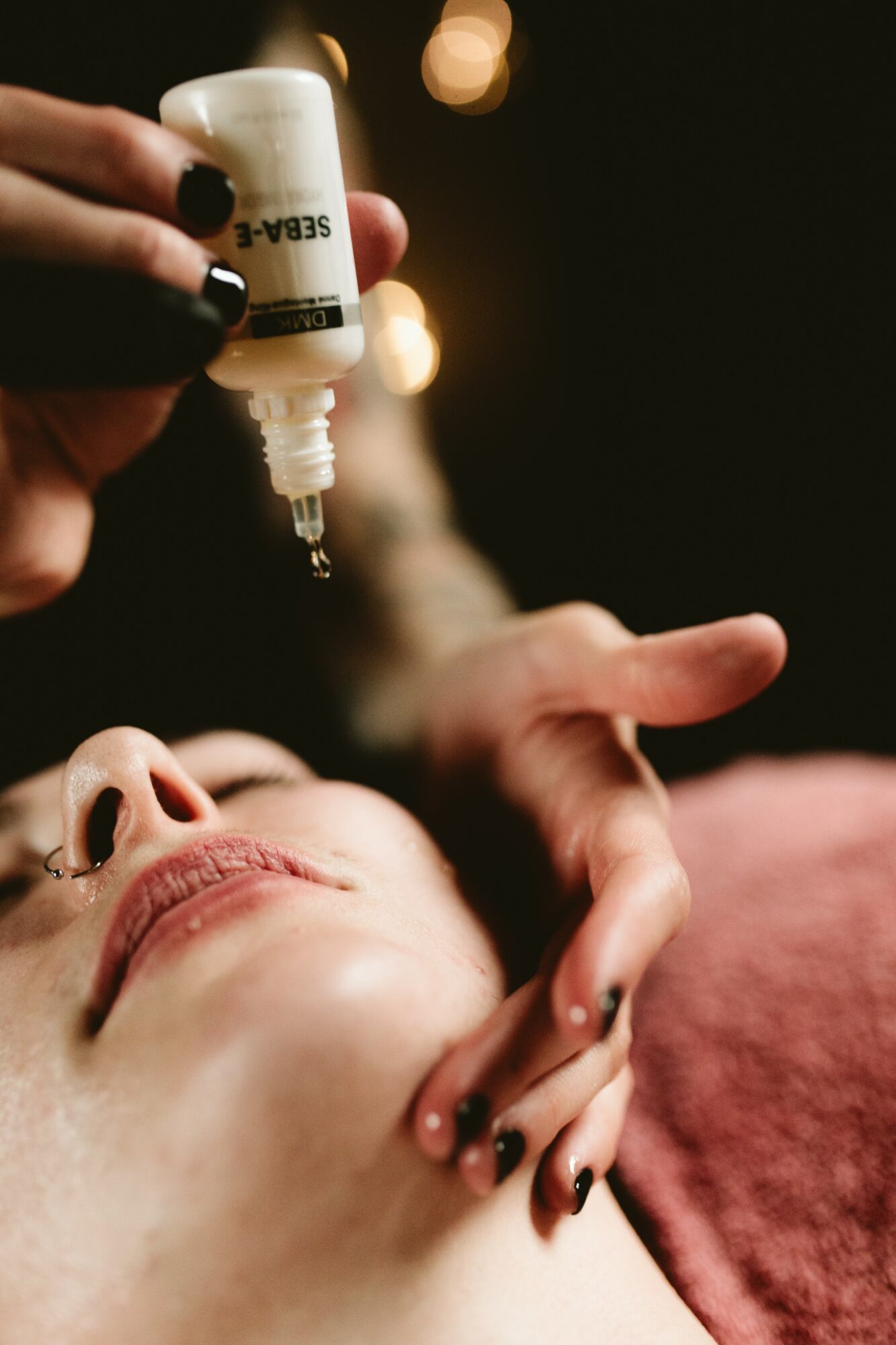Person receiving facial treatment with a dropper, lying down with eyes closed, in a dimly lit setting.