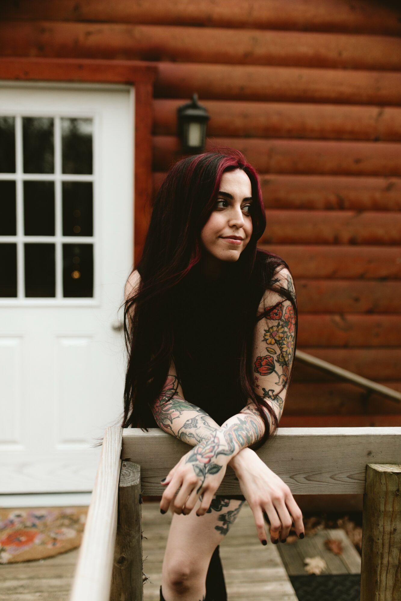 Young woman with long dark hair and tattoos on arms and legs, standing outdoors near a wooden building with a white door.