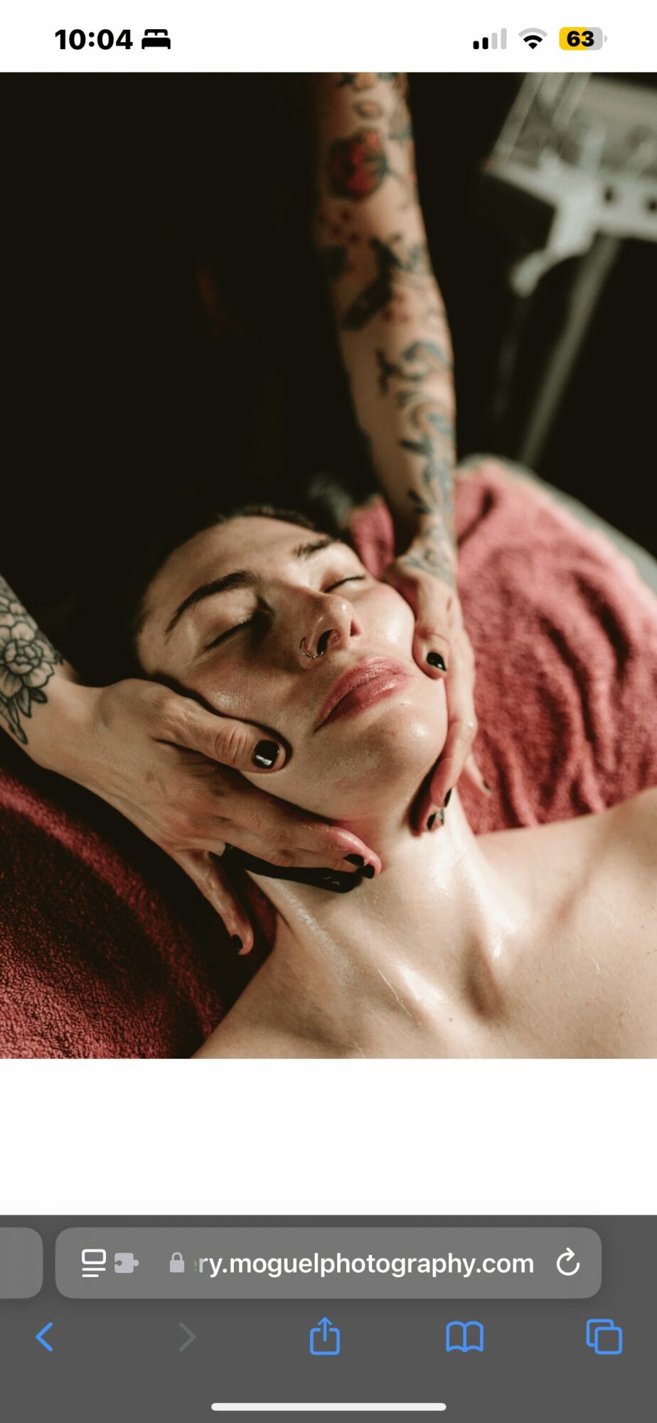 Person receiving a facial massage with eyes closed, lying on a pink towel, tattooed arm visible, dark background.