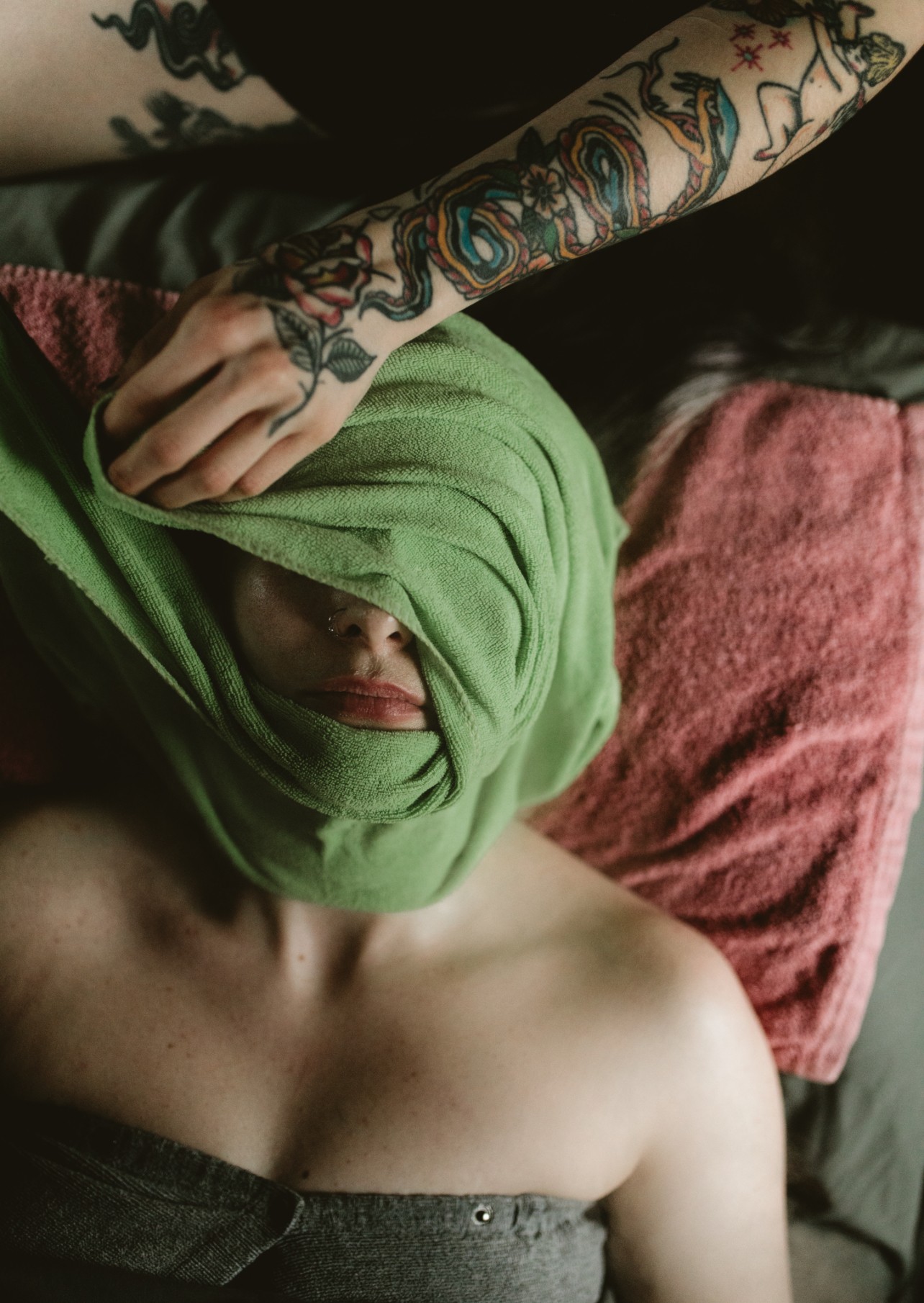 Person lying down with face partially covered by a green cloth, arm with tattoos resting on head, red pillow nearby.