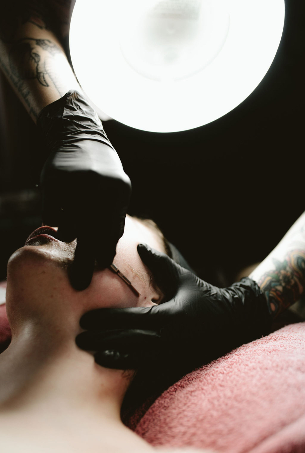Person receiving a tattoo on their neck, with a tattoo artist wearing black gloves working on them, illuminated by a bright light.
