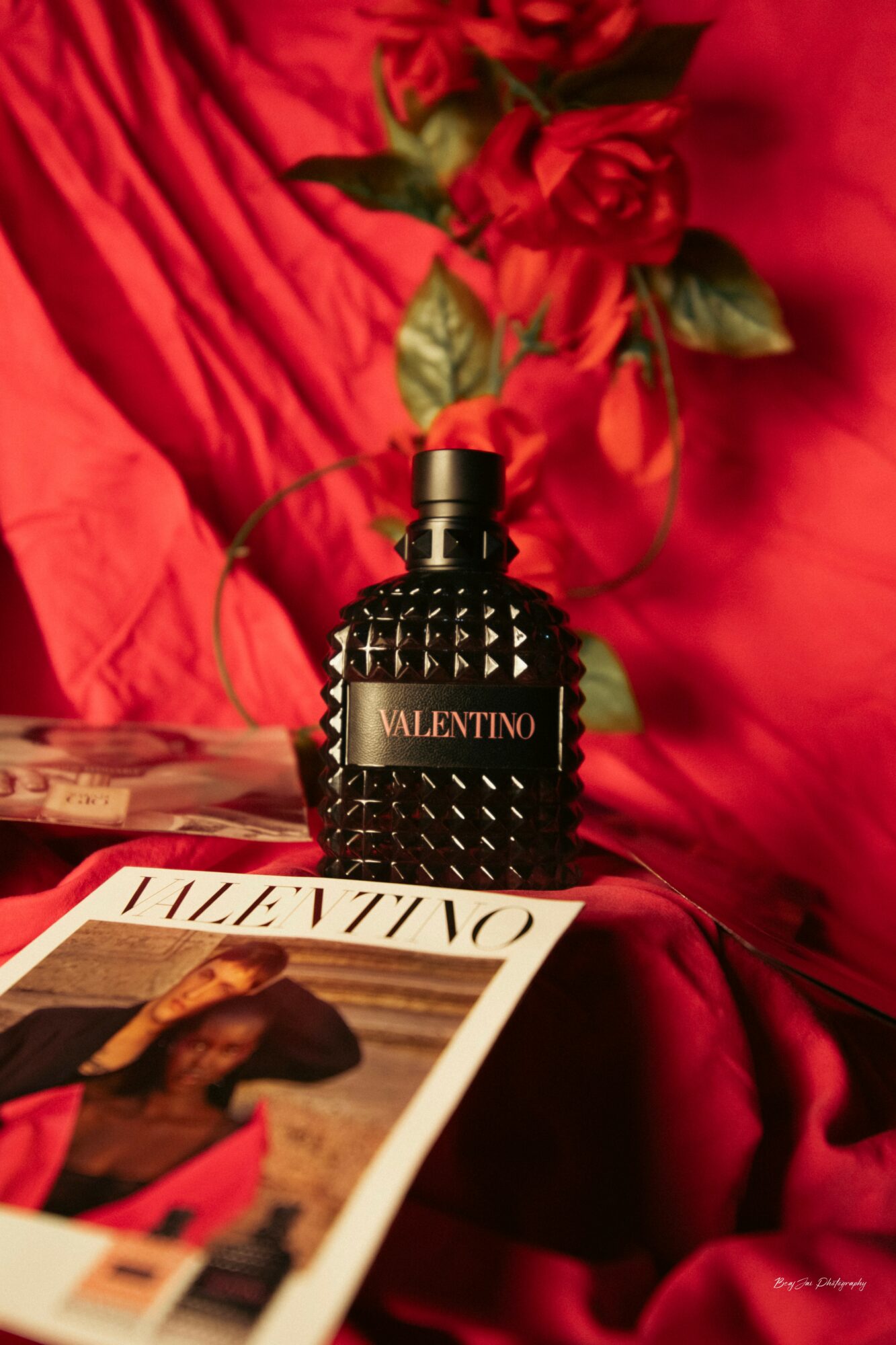 Black Valentino perfume bottle on red fabric with a magazine and a red rose in the background.