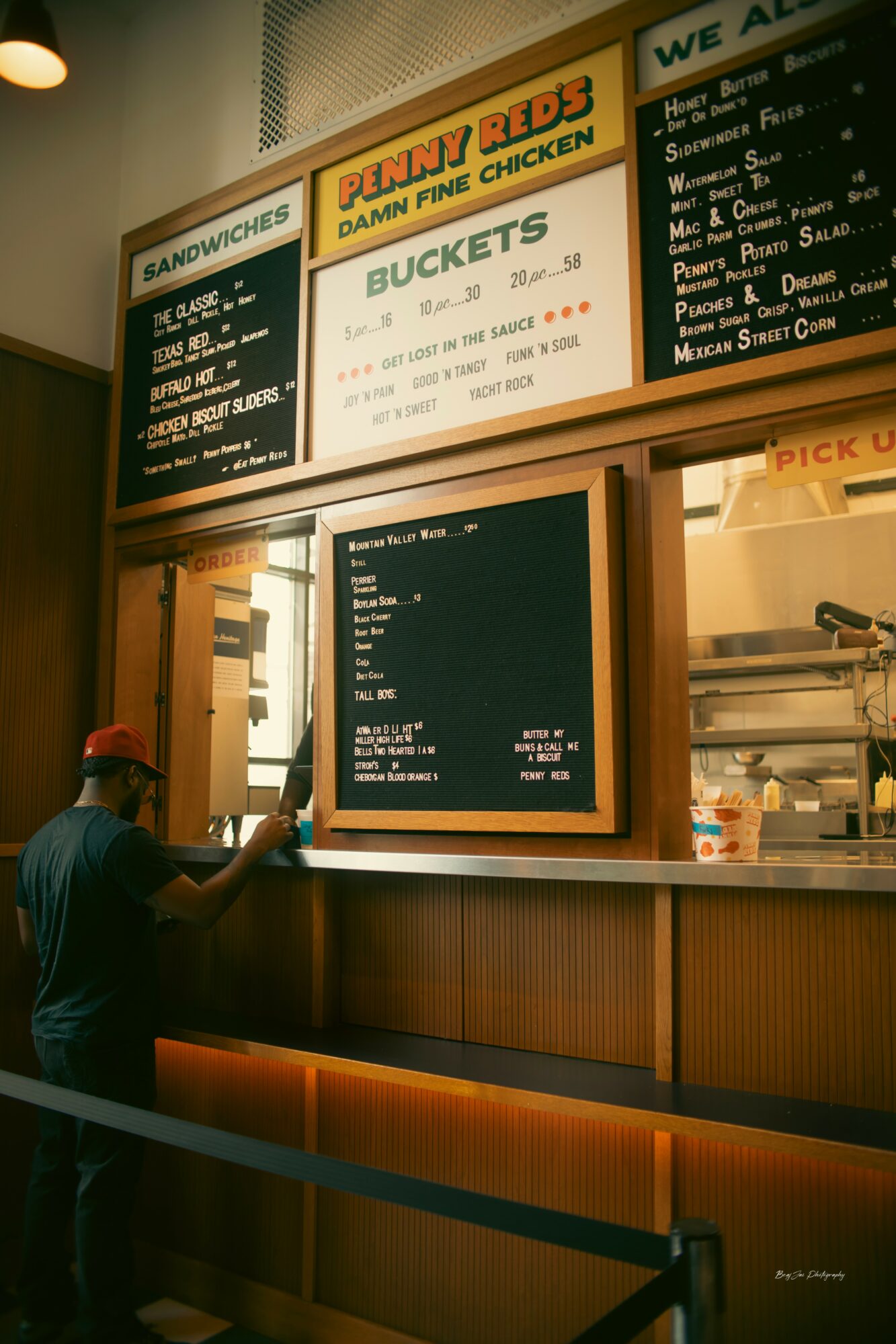 Person in black shirt and red cap writing on a black menu board at a fast-food counter, with illuminated menu signs above.