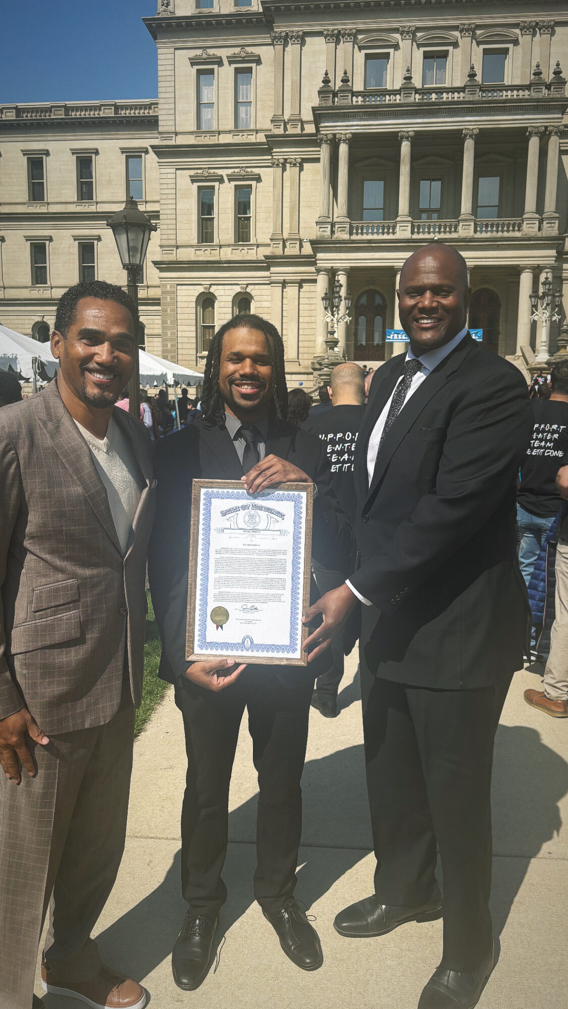 Three men in suits standing outdoors in front of a historic building, smiling, one holding a framed certificate.
