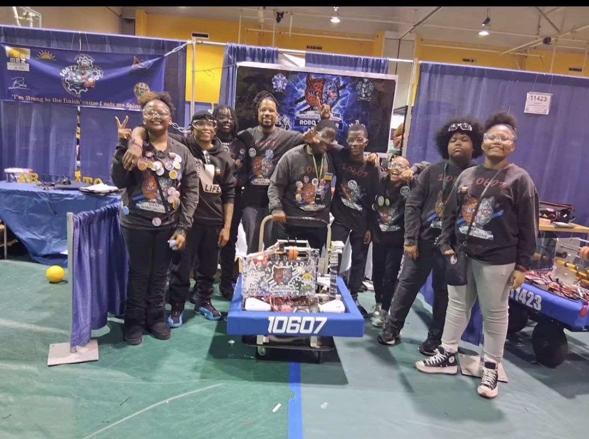 Group of eight people standing behind a robot with the number 10607 at an indoor event, with blue curtains and display tables in the background.