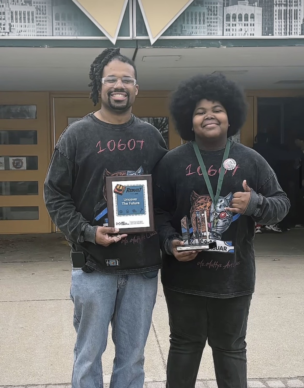 Two smiling people holding awards and a microphone, standing outside a building with glass doors.