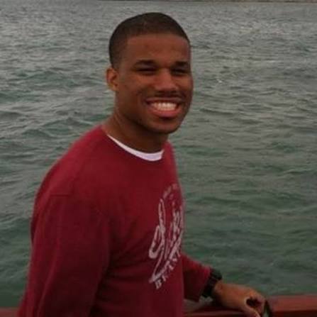 Young man smiling outdoors near water, wearing a red shirt, with a background of water and sky.